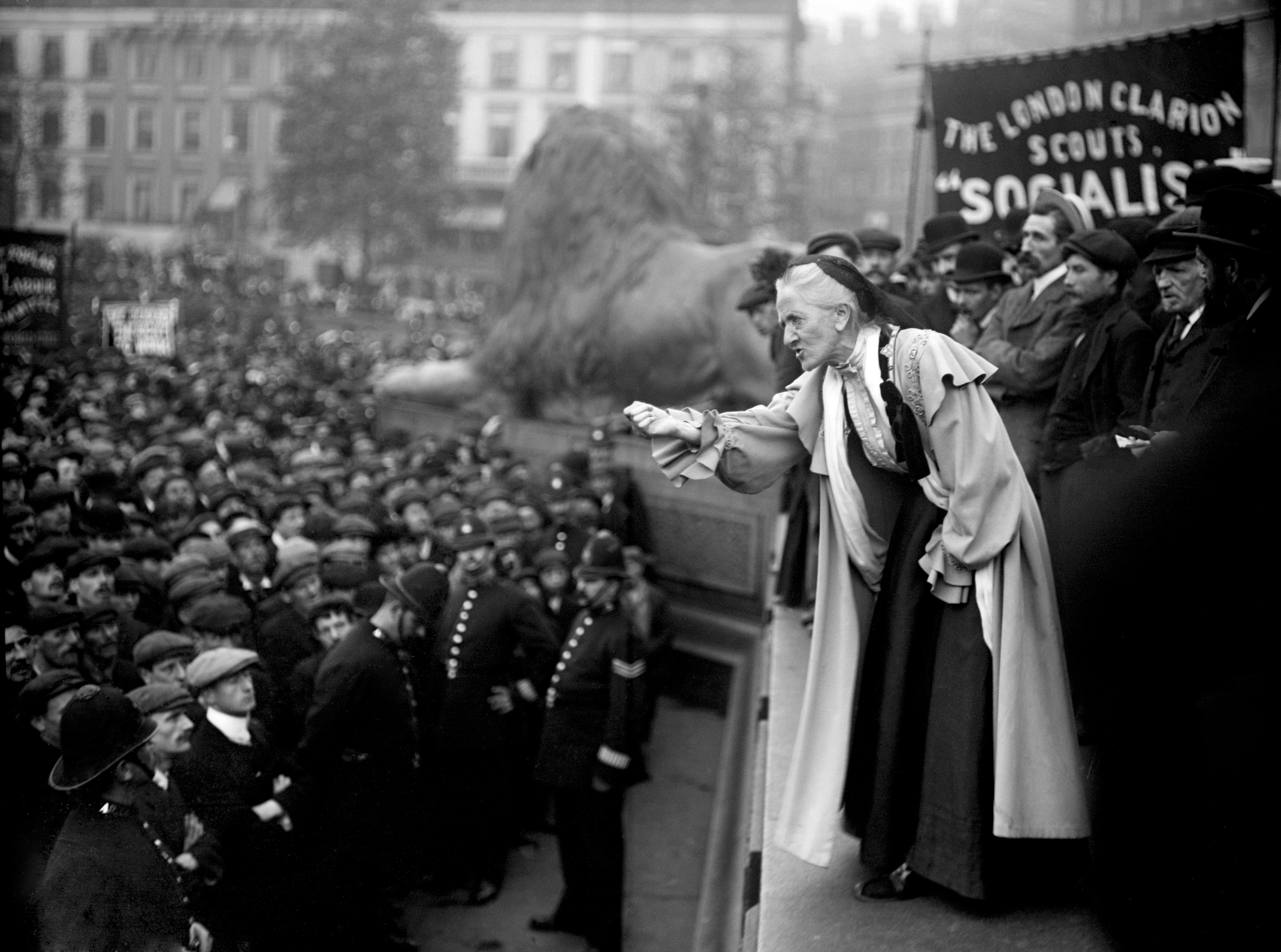 An older woman speaks to male and female crowd with lion statues in the background
