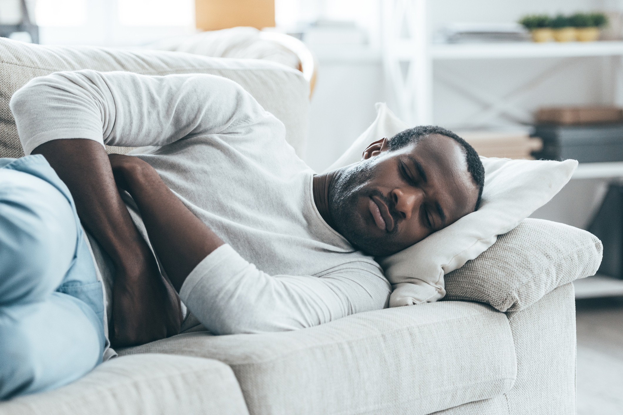 Handsome young African man keeping eyes closed while lying on the sofa and touching his stomach