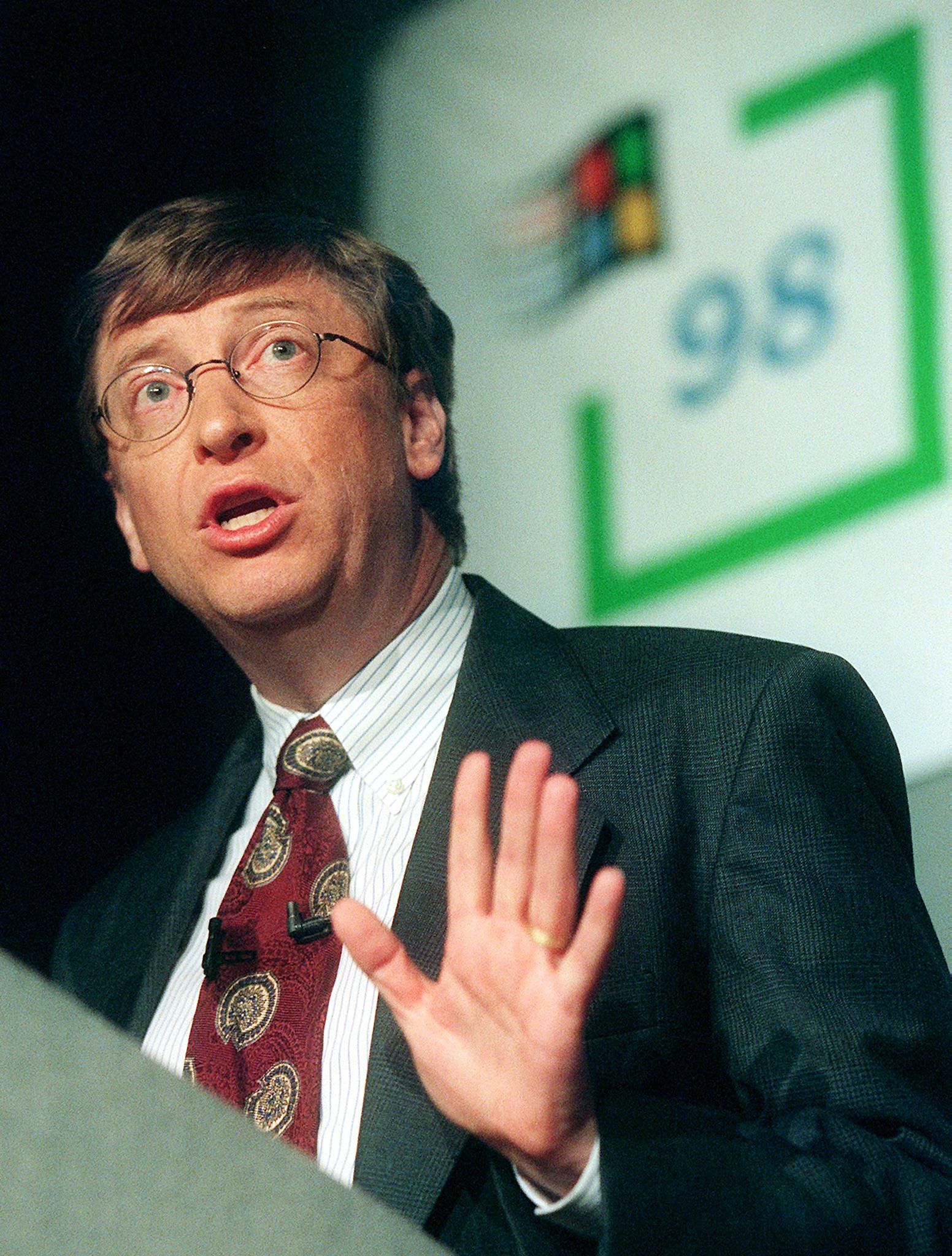 A younger Gates speaks at a platform with the Windows 98 logo behind him