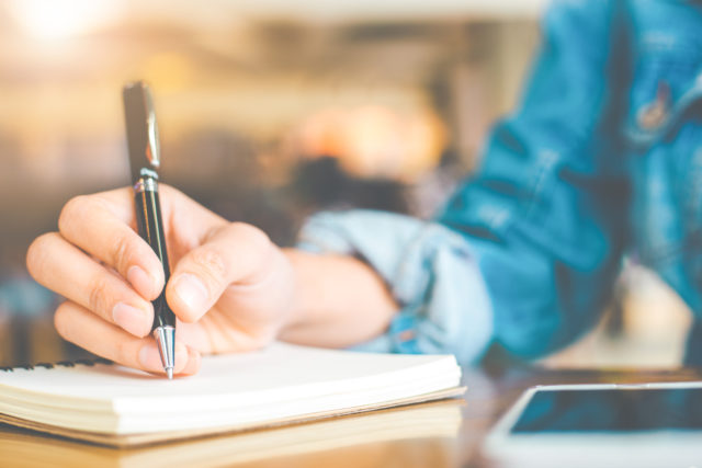 Woman's hand writing on a notepad with a pen on a wooden desk.