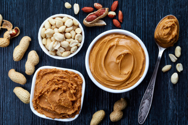 Two bowls of peanut butter and peanuts on dark wooden background from top view