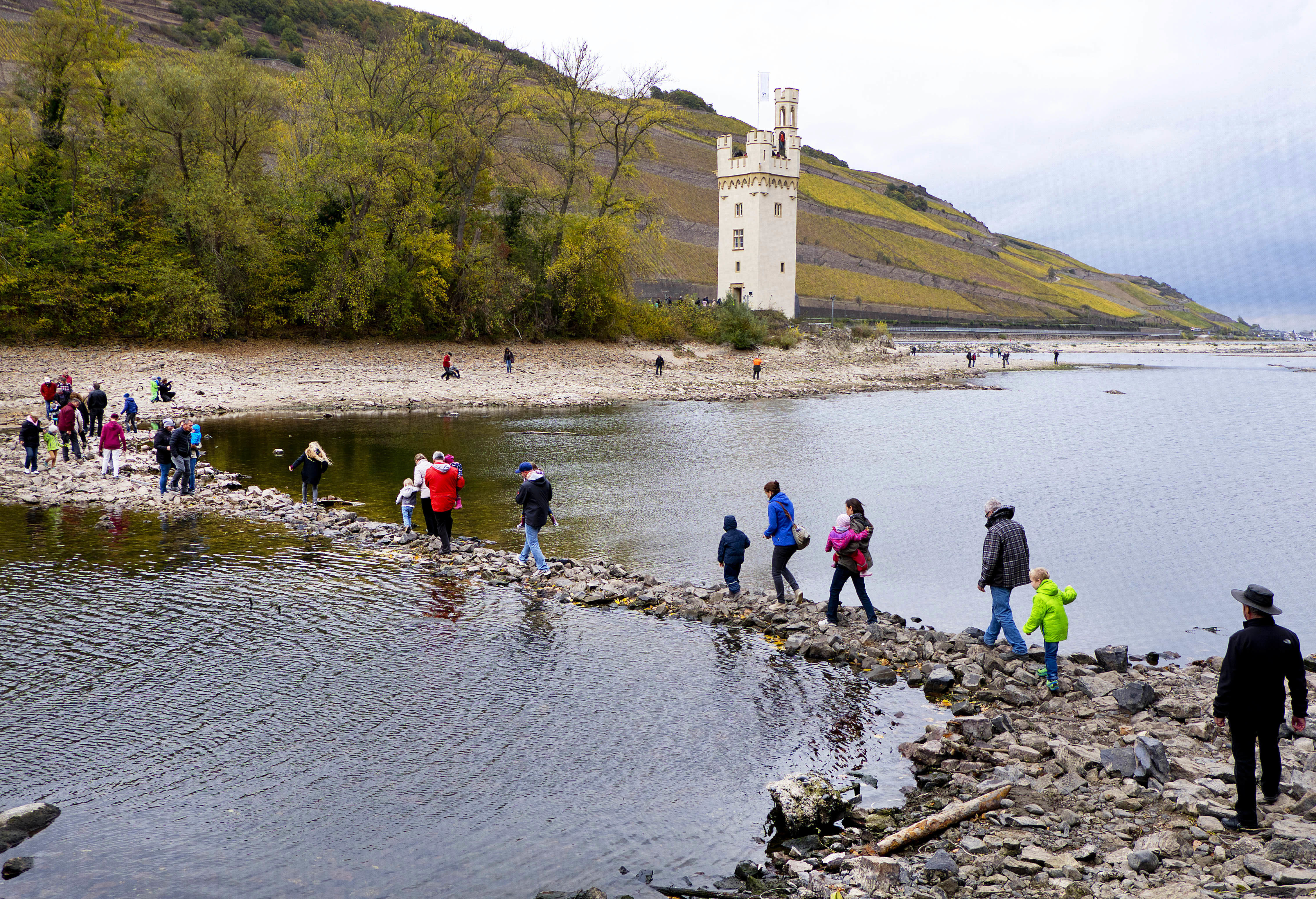People walk over stones looking out of the River Rhine to the Maeuseturm (mice tower) of Bingen