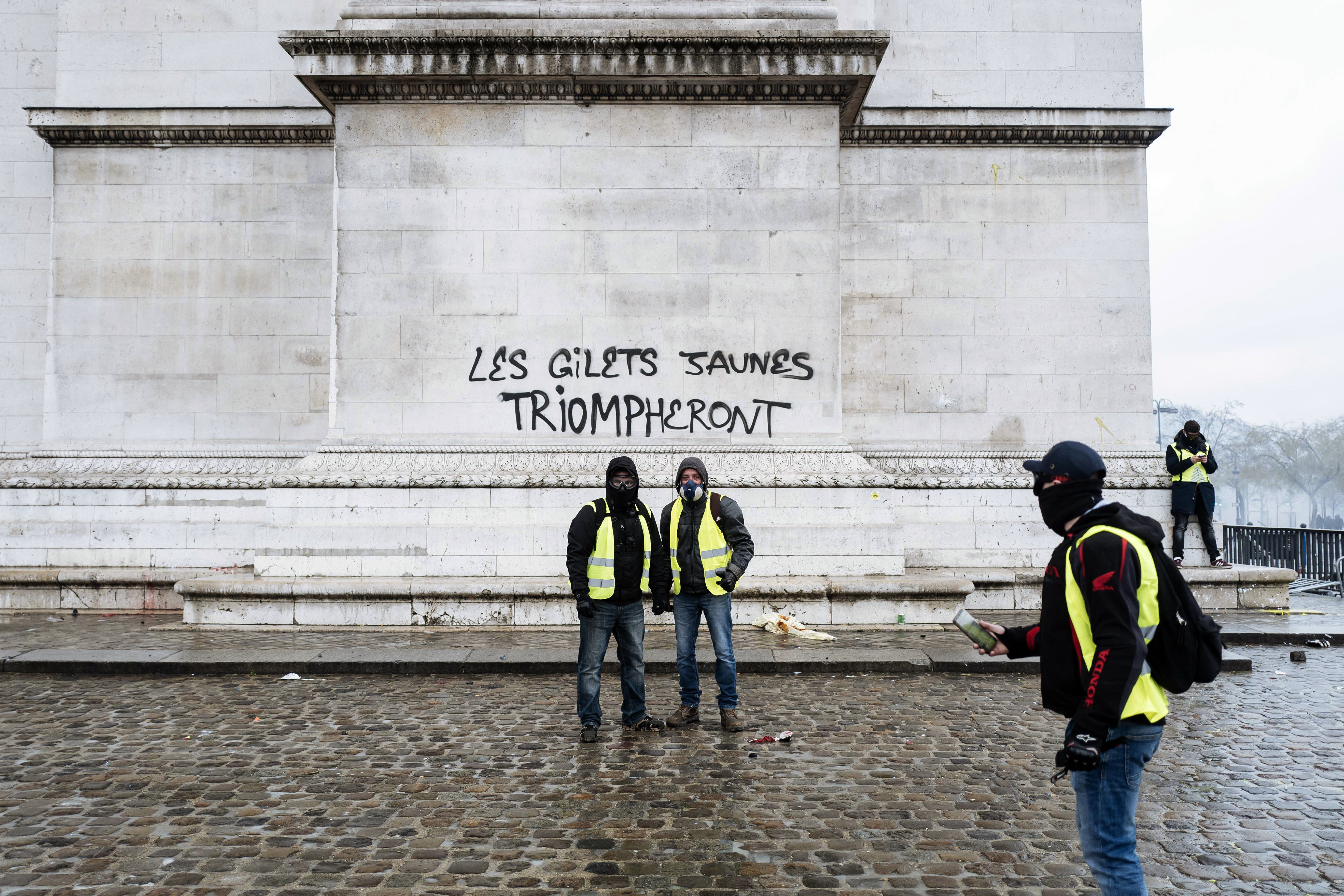 Demonstrators stand by the words 'The yellow jackets will triumph' written in big black letters at the base of the Arc de Triomphe