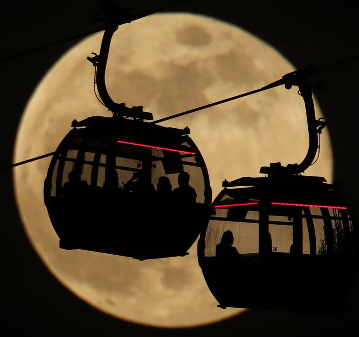 Emirates Air Line cable cars are silhouetted against the backdrop of the moon in Greenwich, London
