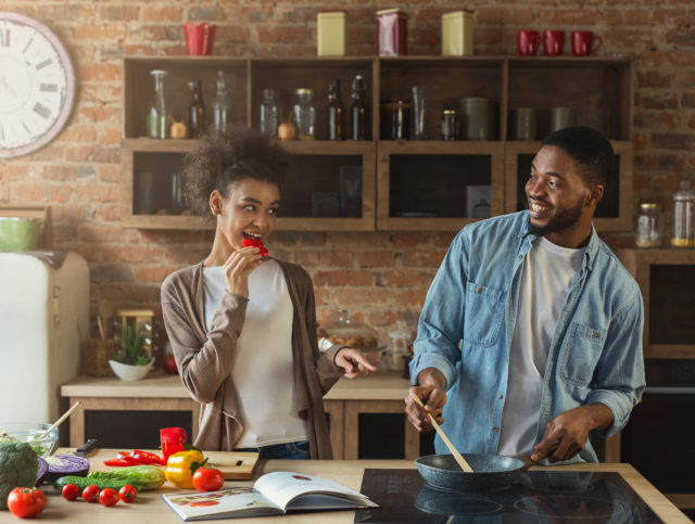 Happy black couple preparing dinner with recipe together in modern kitchen. Family frying vegetables