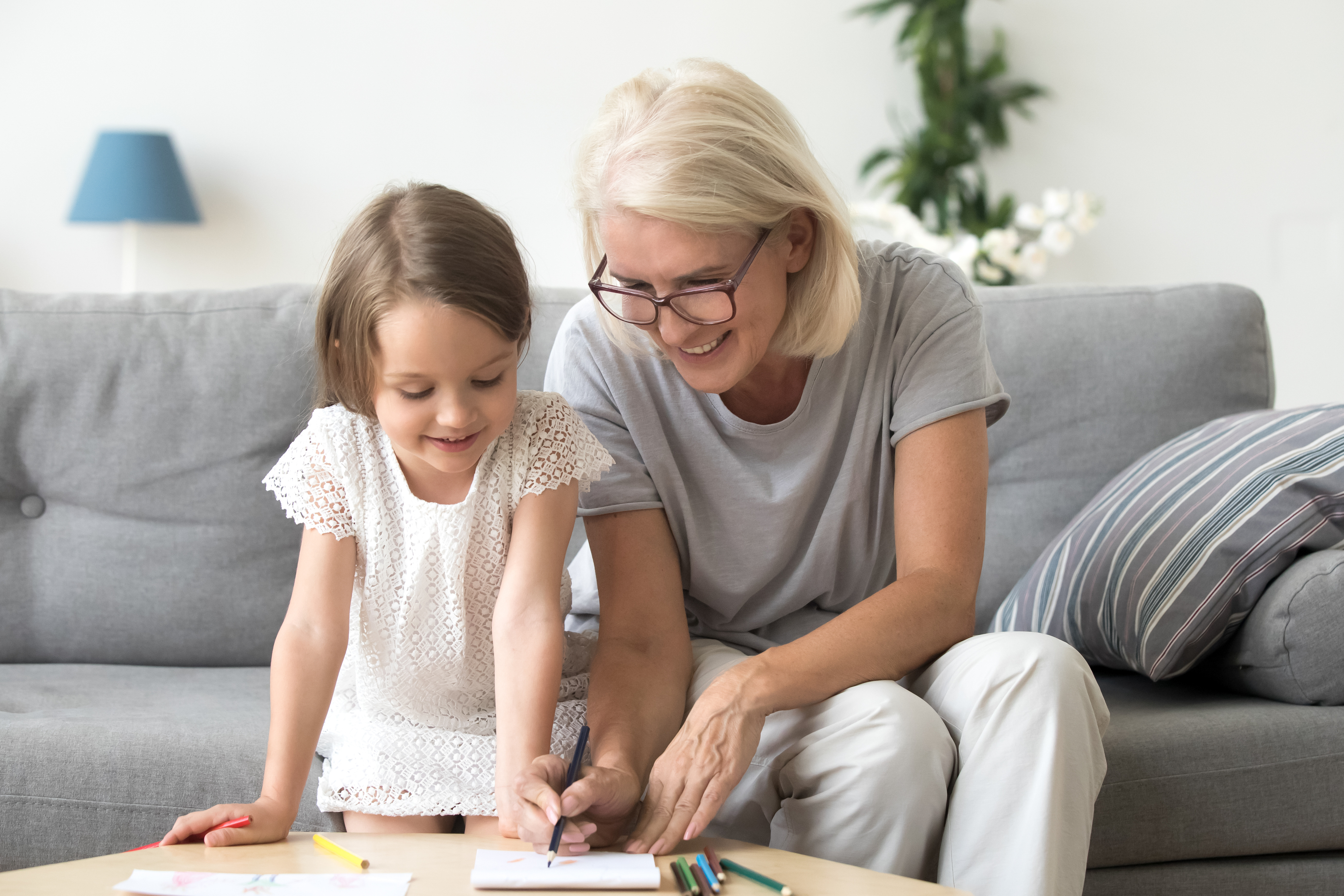 Smiling grandmother and little kid granddaughter drawing on paper with colored pencils together, caring granny teaching grandchild having fun playing at home, grandma and creative child activity