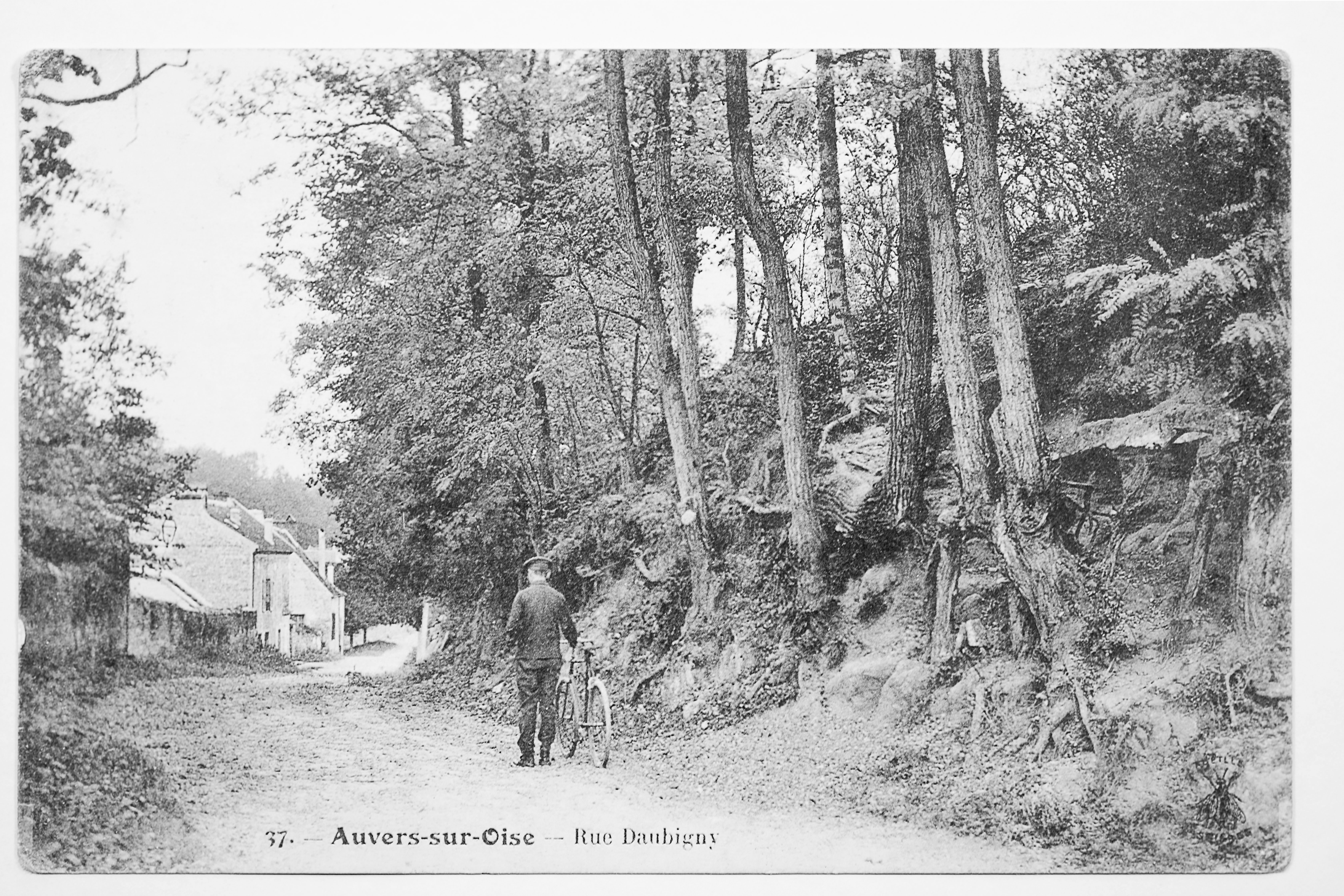 A faded picture postcard featuring a man standing next to a bicycle on a back street of the village of Auvers-sur-Oise