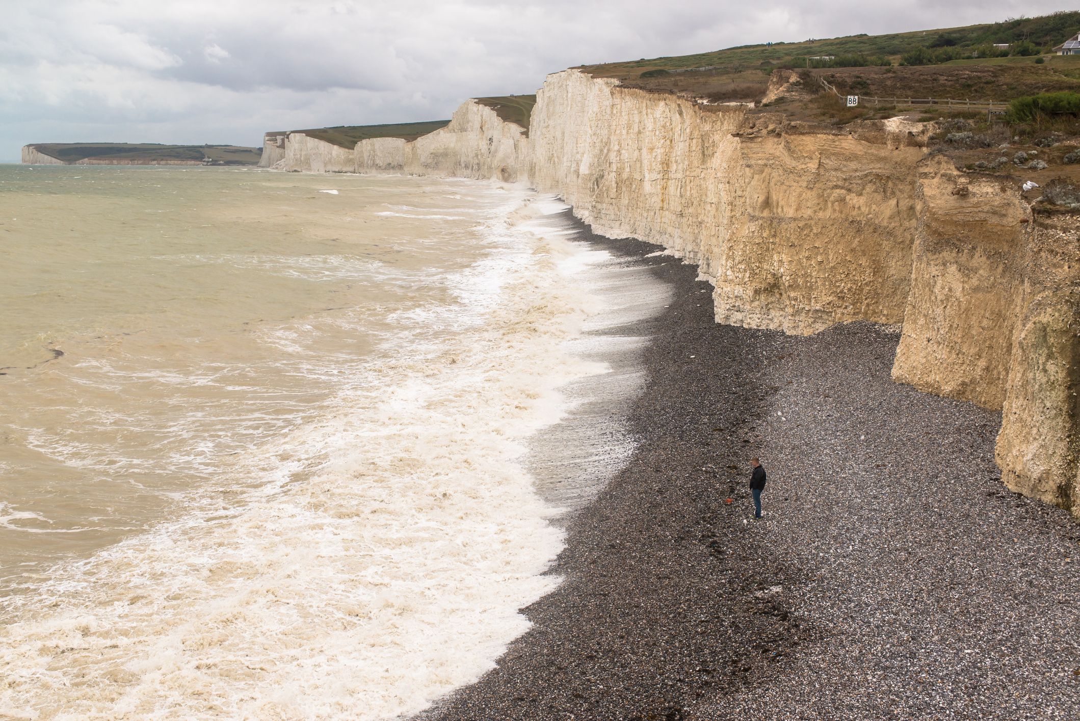 Solitary man standing alone on a gravel beach below the white cliffs of Seven Sisters facing rough sea and waves.