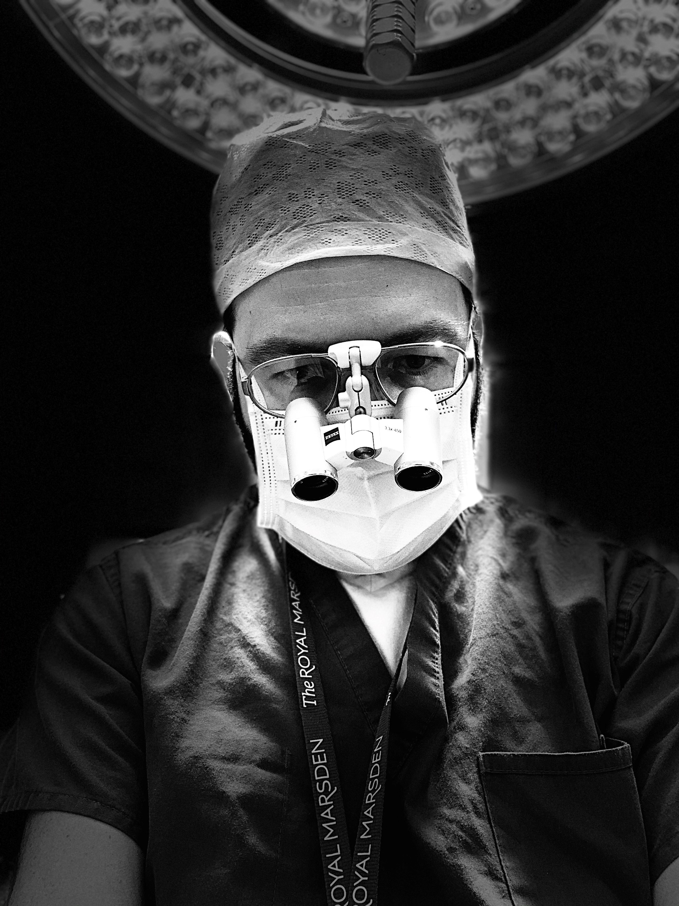 A black and white photo of a surgeon in an operating theatre. He is weirding a mask and glasses,