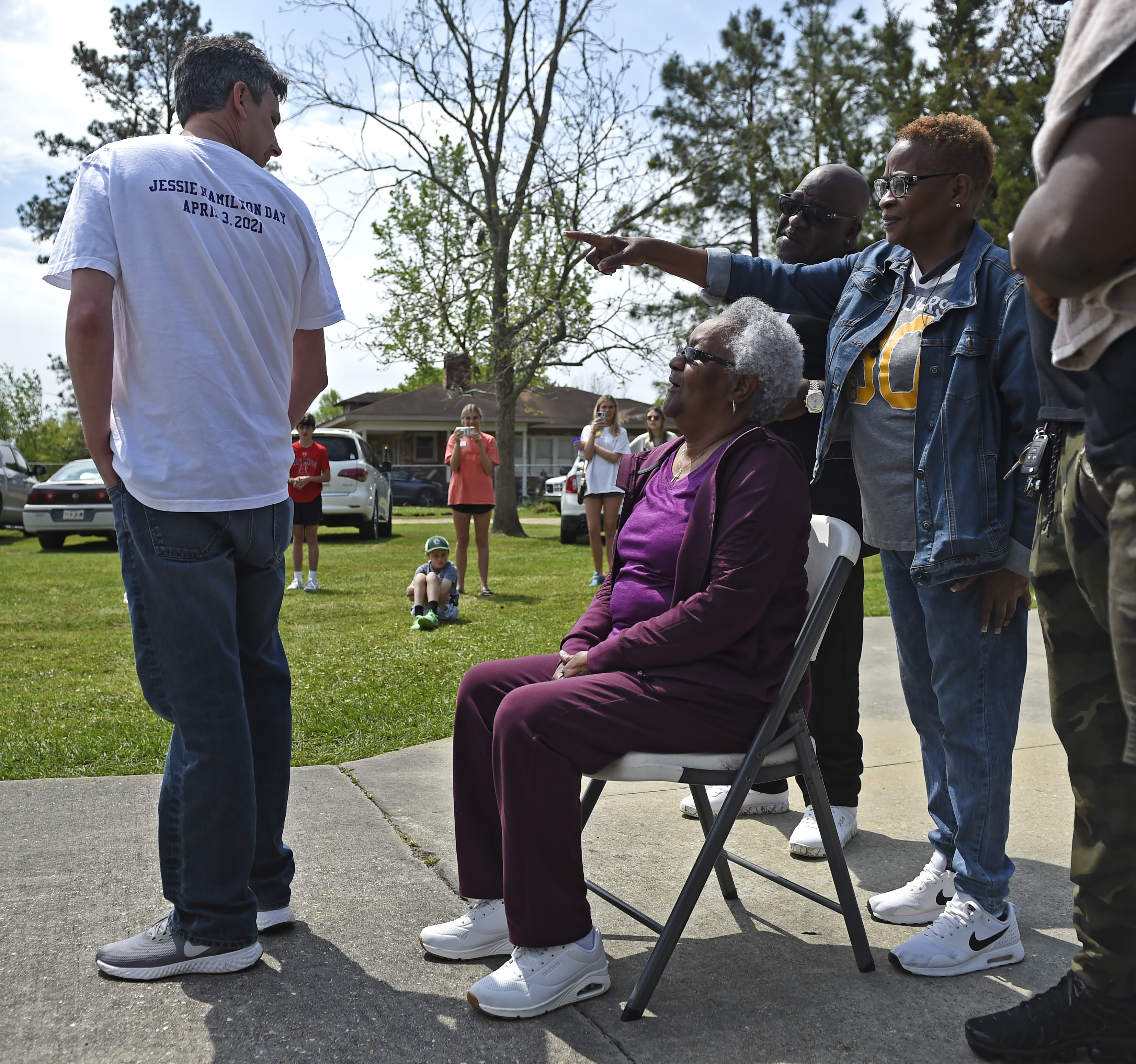 Andrew Fusaiotti, left, turns around to show Jessie Hamilton, seated, the back of his shirt as Ms Hamilton's daughter, Yonetta Tircuit, right, points as LSU FIJI graduates gather to surprise their former house kitchen staff member to pay off her mortgage and celebrate Jessie Hamilton Day in Baker, Louisiana