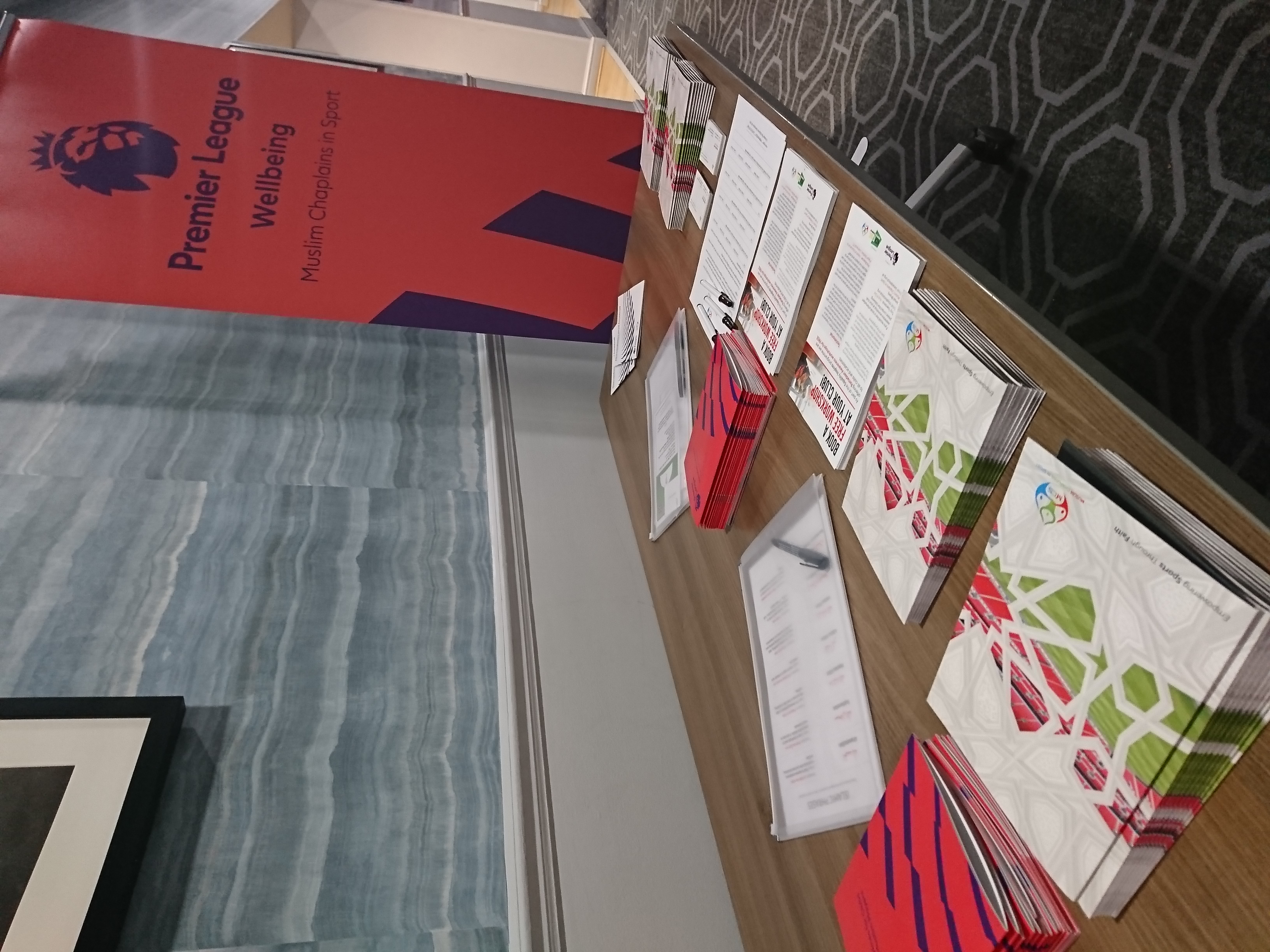 A picture of some leaflets and literature from the Muslim Chaplains In Sport, on a light brown table with an orange Premier League sign next to it that reads 'Premier League Wellbeing - Muslim Chaplains in Sport'