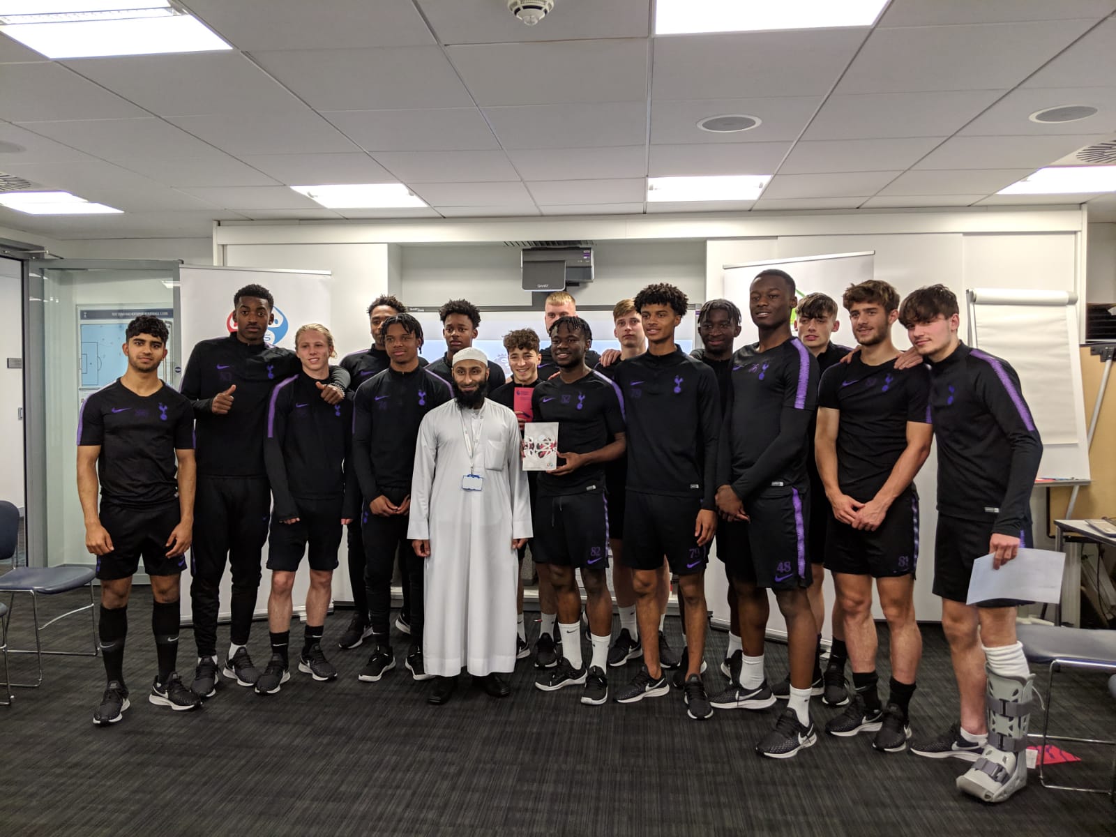 Ismail Bhamji stands in white clothing in front of a number of Tottenham footballers on a grey carpet in a lit room