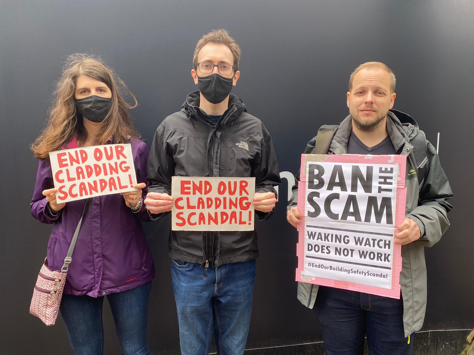 A woman in a purple coat and a black mask holds a white sign with red writing, as does a man in a black jacket and a black face mask with glasses, while a man in a grey jacket holds a white sign with black writing