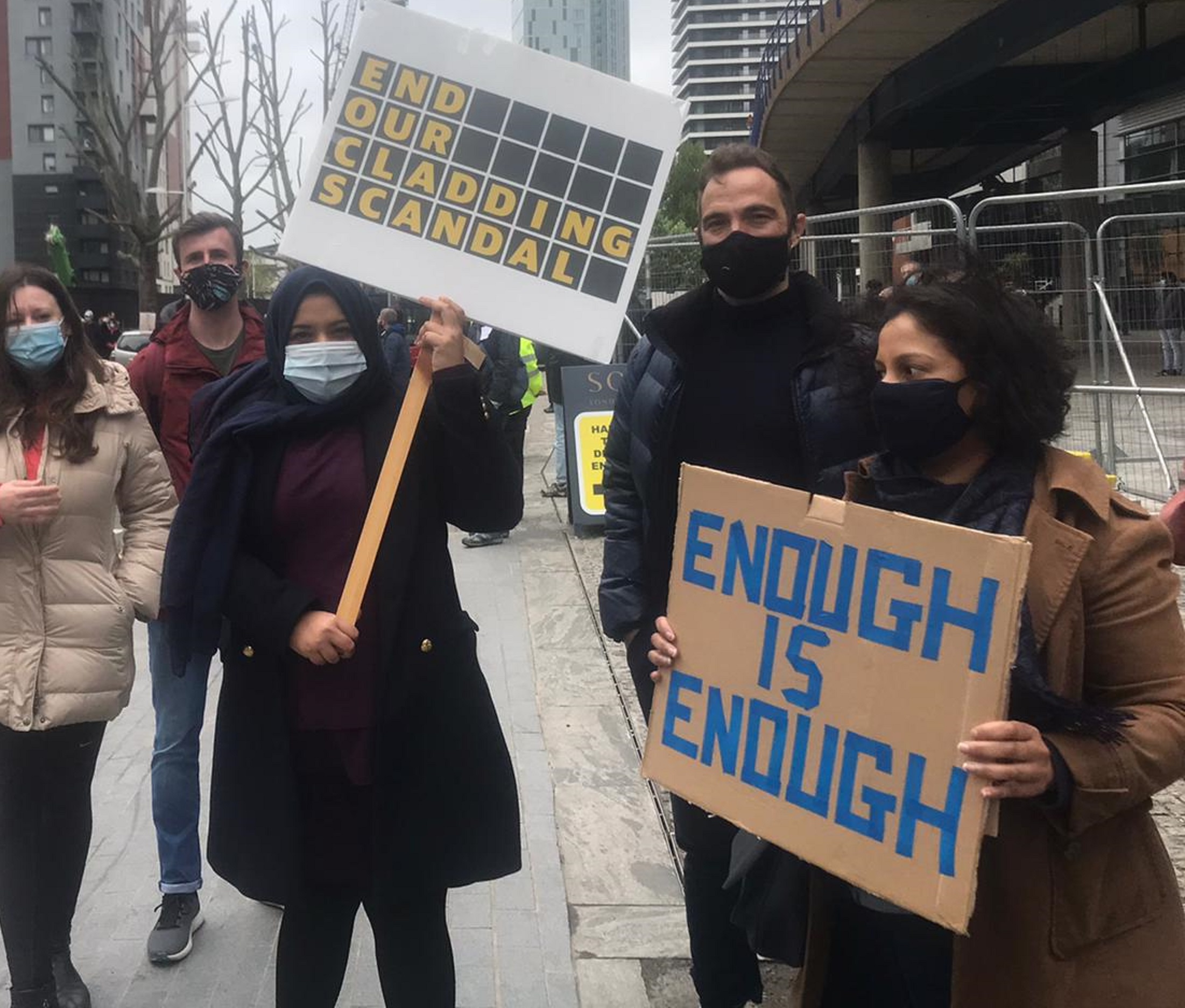 Signs held by protesters, which read 'END OUR CLADDING SCANDAL' and 'ENOUGH IS ENOUGH' - A woman with her head covered, wearing a light blue mask, holds a sign with yellow writing on a black background. To her right, a woman wearing a black mask and a brown coat holds a cardboard sign with blue lettering