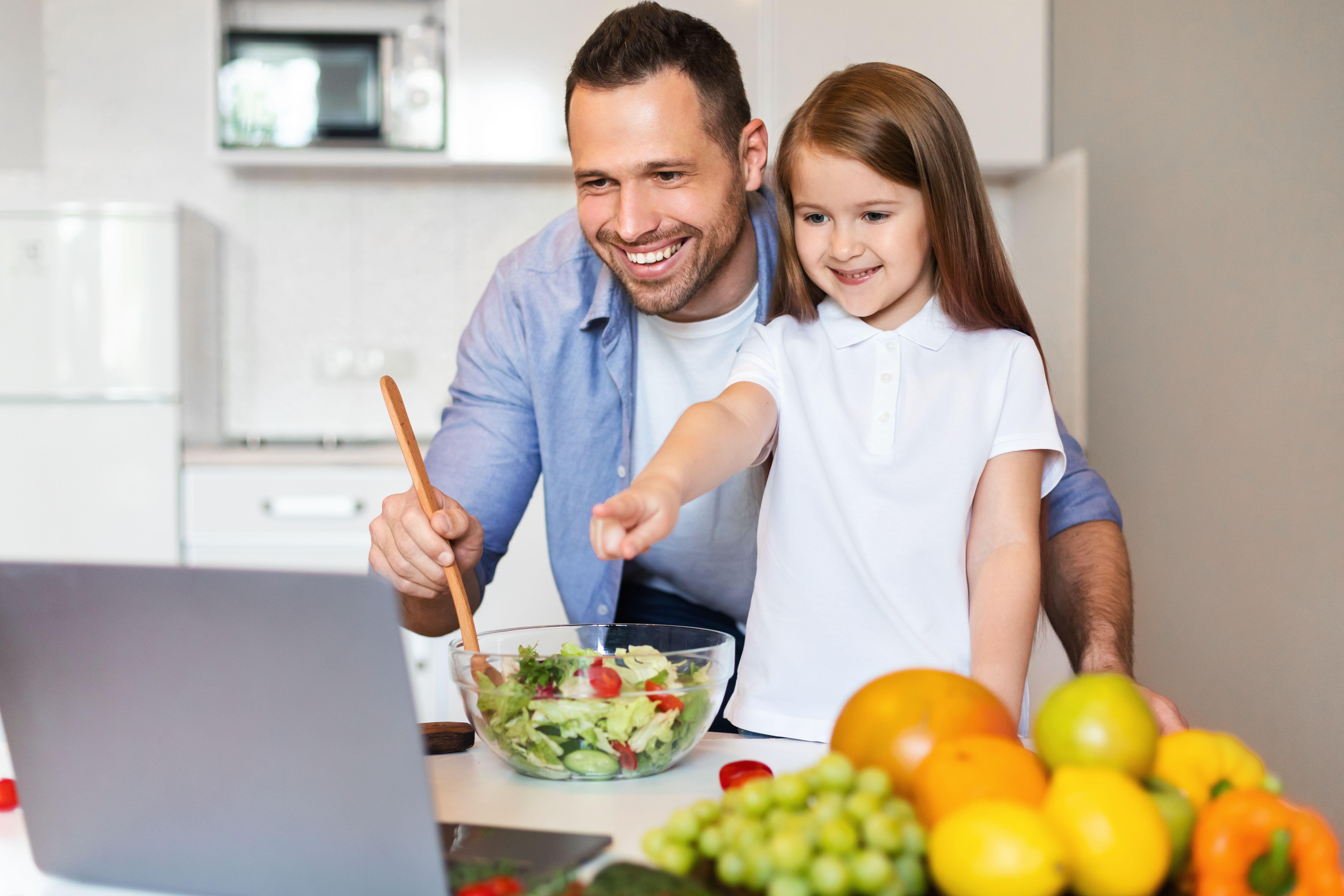 Cute Father And Daughter Cooking Using Laptop Together In Kitchen