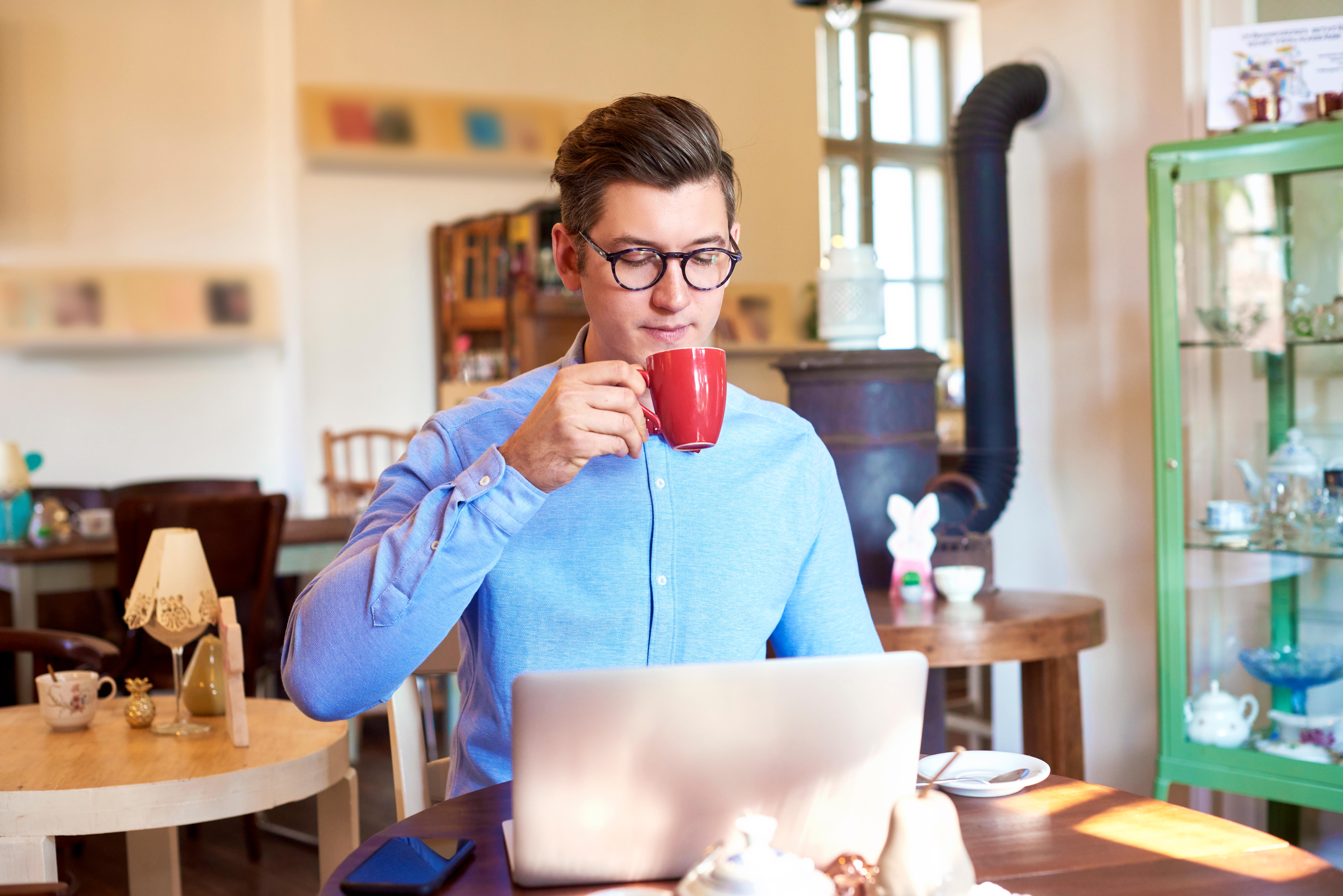 Young man having coffee while sitting at table and using his laptop in small cafe