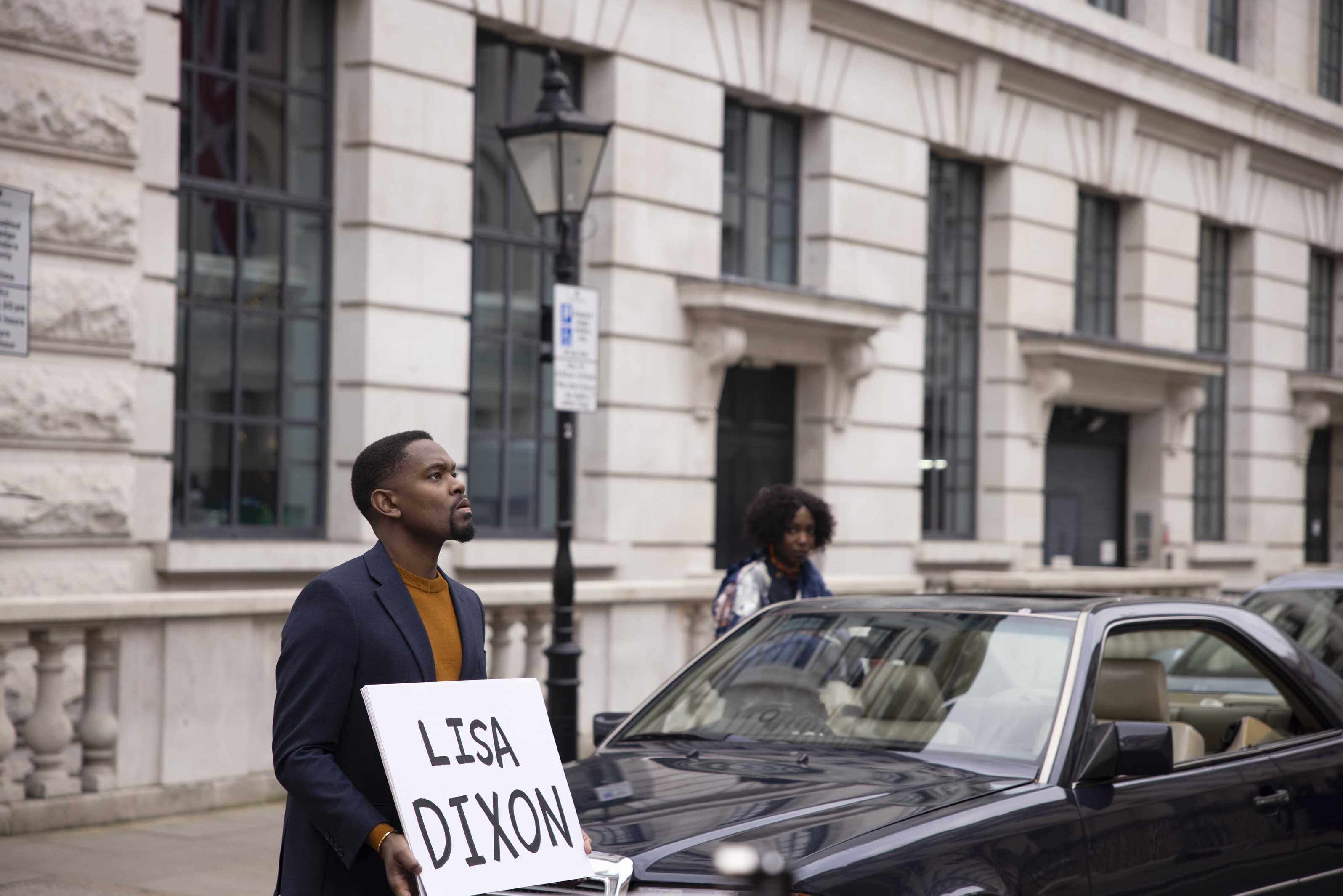 Aml Ameen as Melvin in a scene from Boxing Day, standing in the street holding a sign that reads 'Lisa Dixon'