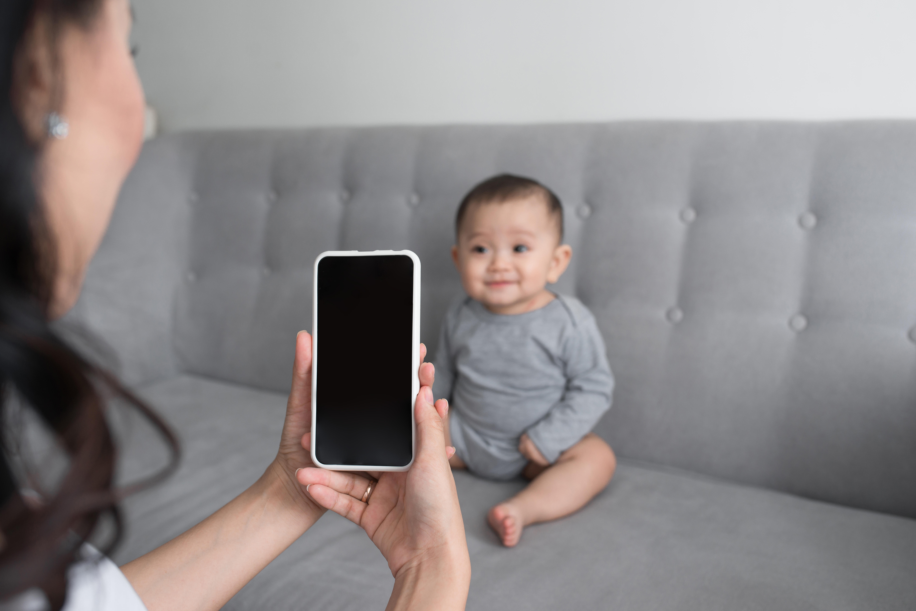 mother taking a picture of her baby girl on sofa with smartphone