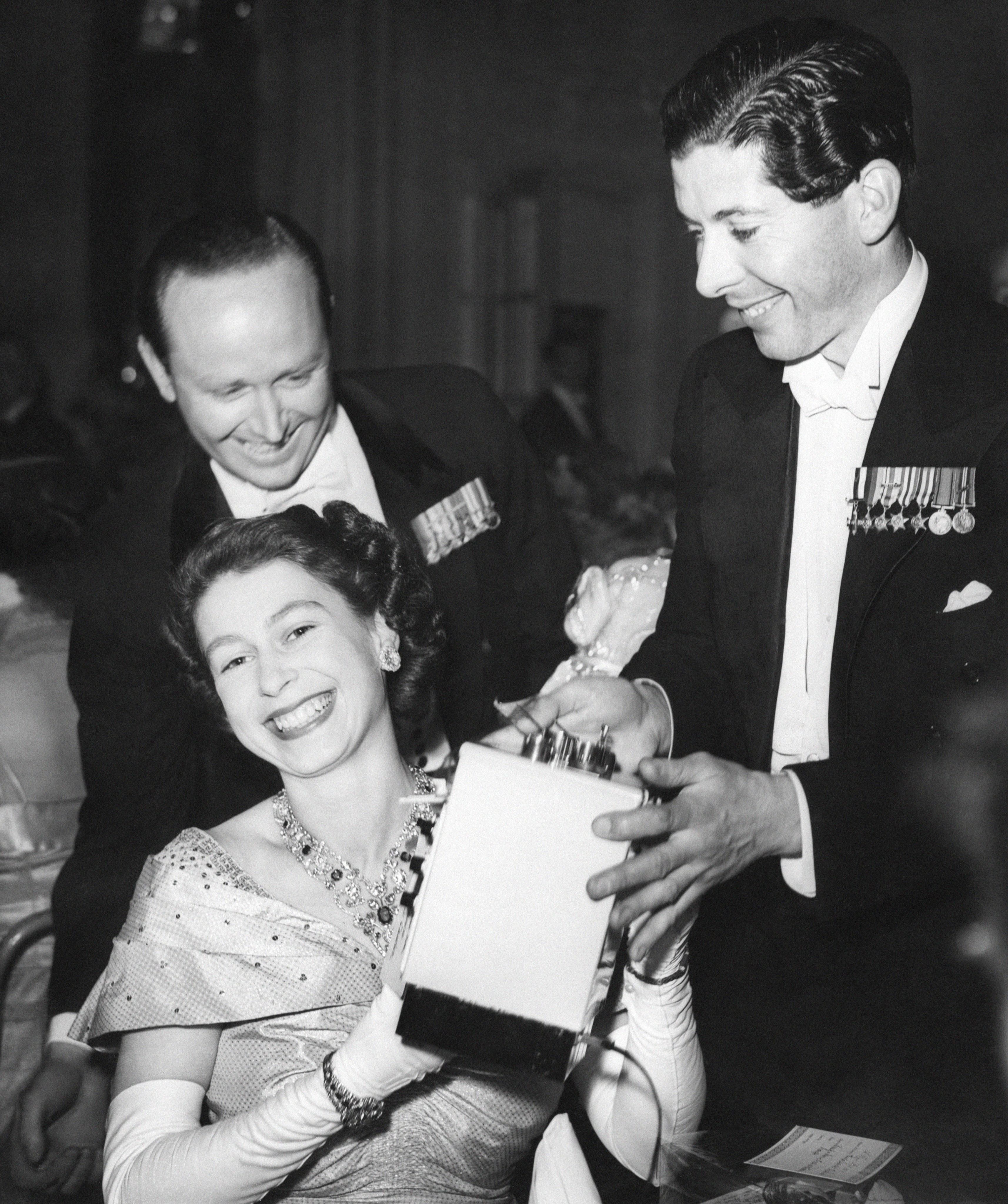 A laughing Princess Elizabeth accepts a toy stove for Prince Charles from Lieutenant Michael Parker, R.N. left, Equerry-in-Waiting to the Princess and the Duke of Edinburgh, and the Hon. Piers St. Aubyn at the Savoy Hotel, London. The Princess was at the Flower Ball in aid of St. Loyes College for the the Training and Rehabilitation of the Disabled