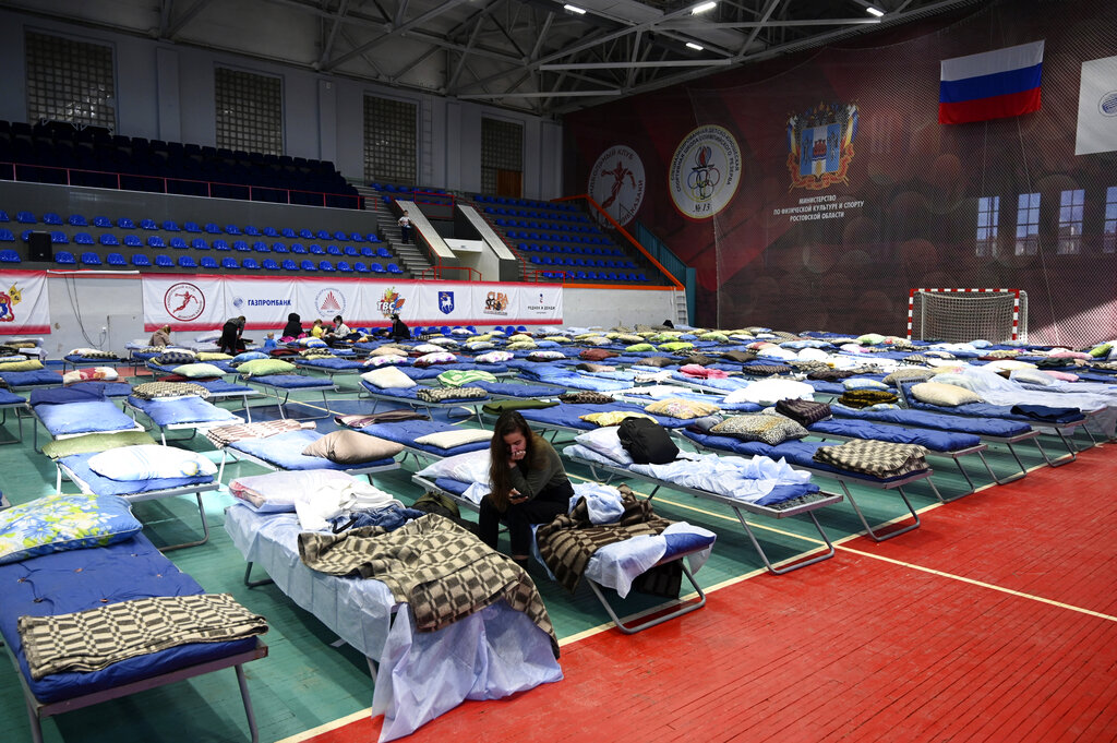 Displaced citizens sit on camp beds in large sporting hall.