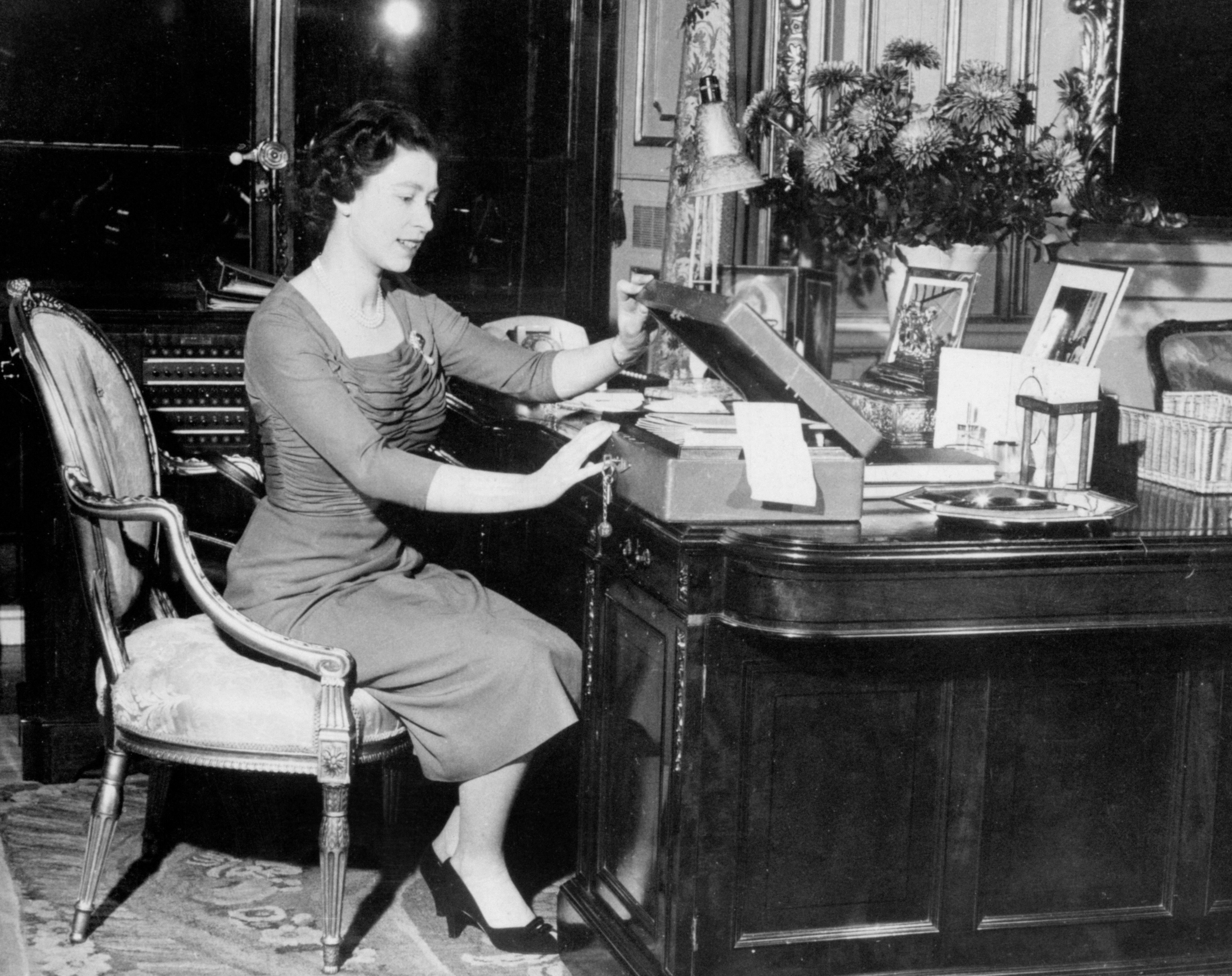 Queen Elizabeth II at work, seated at her desk in Buckingham Palace, London. The Queen is seen opening one of the 'boxes' in which documents and papers, sorted for her attention, are sent upstairs by the Private Secretary. Behind the Queen is the Palace switchboard.