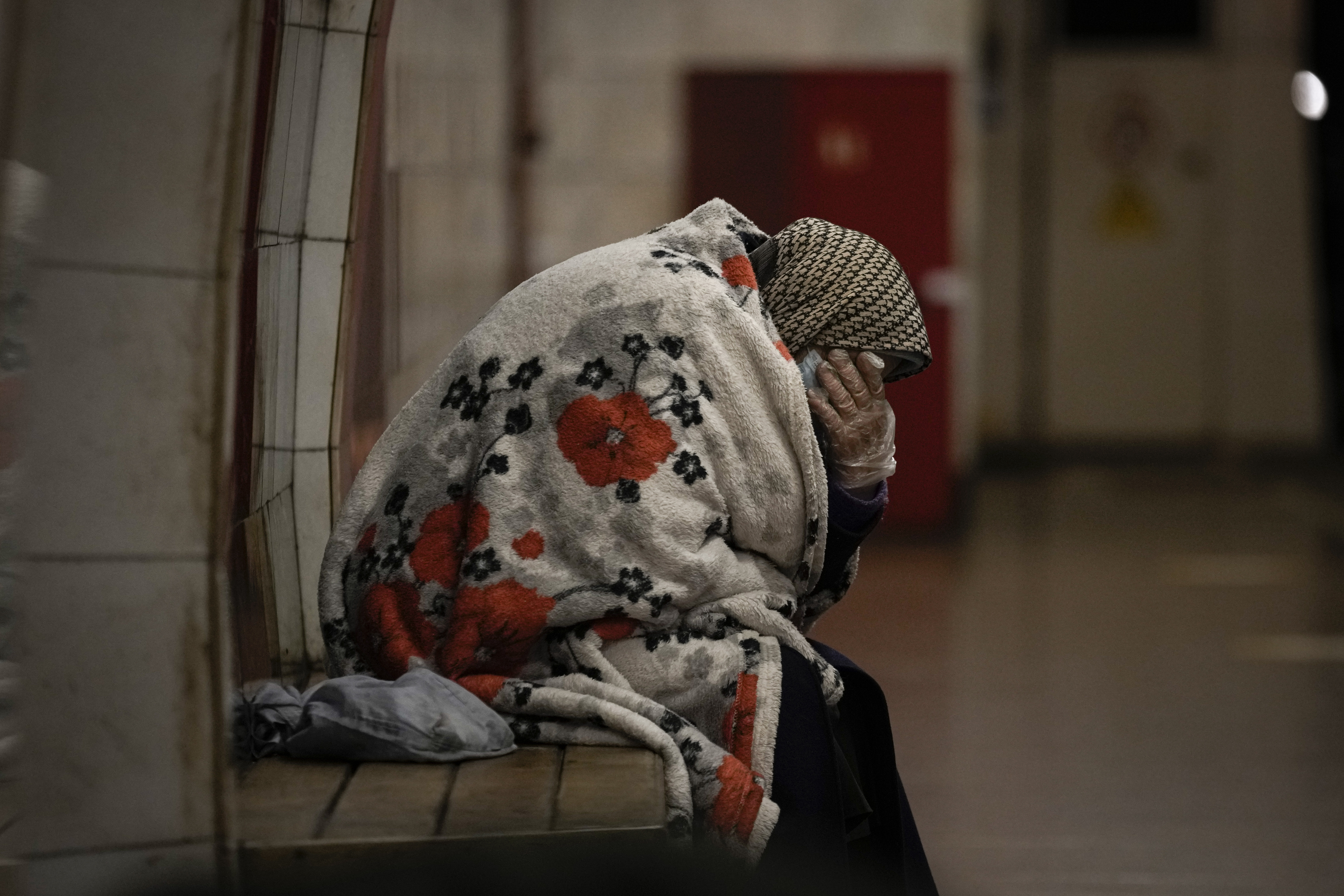 An elderly woman sits on a bench wrapped in a blanket in a subway station turned into a shelter in Kyiv, Ukraine, 