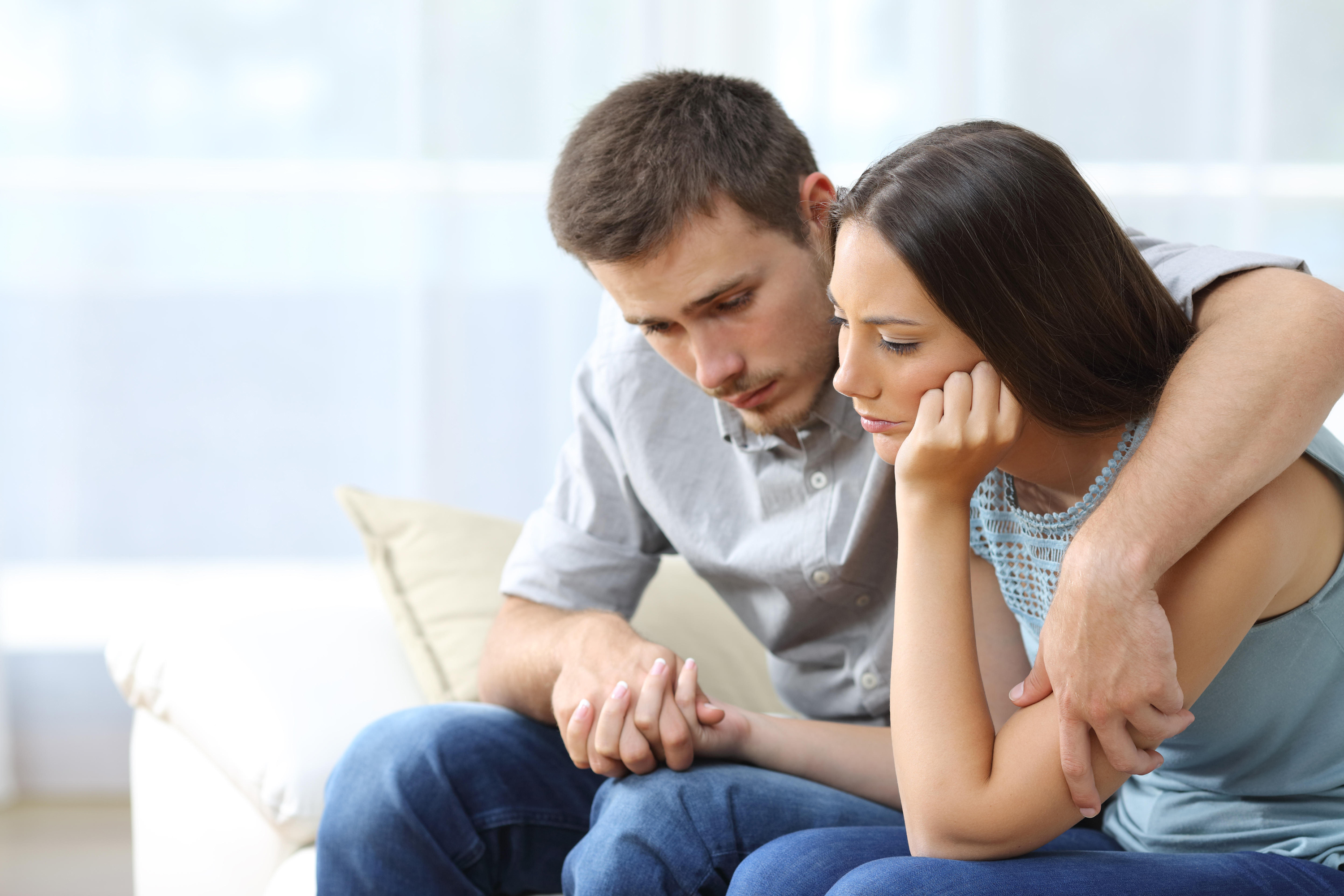 Sad couple comforting each other sitting on a couch in the living room at home
