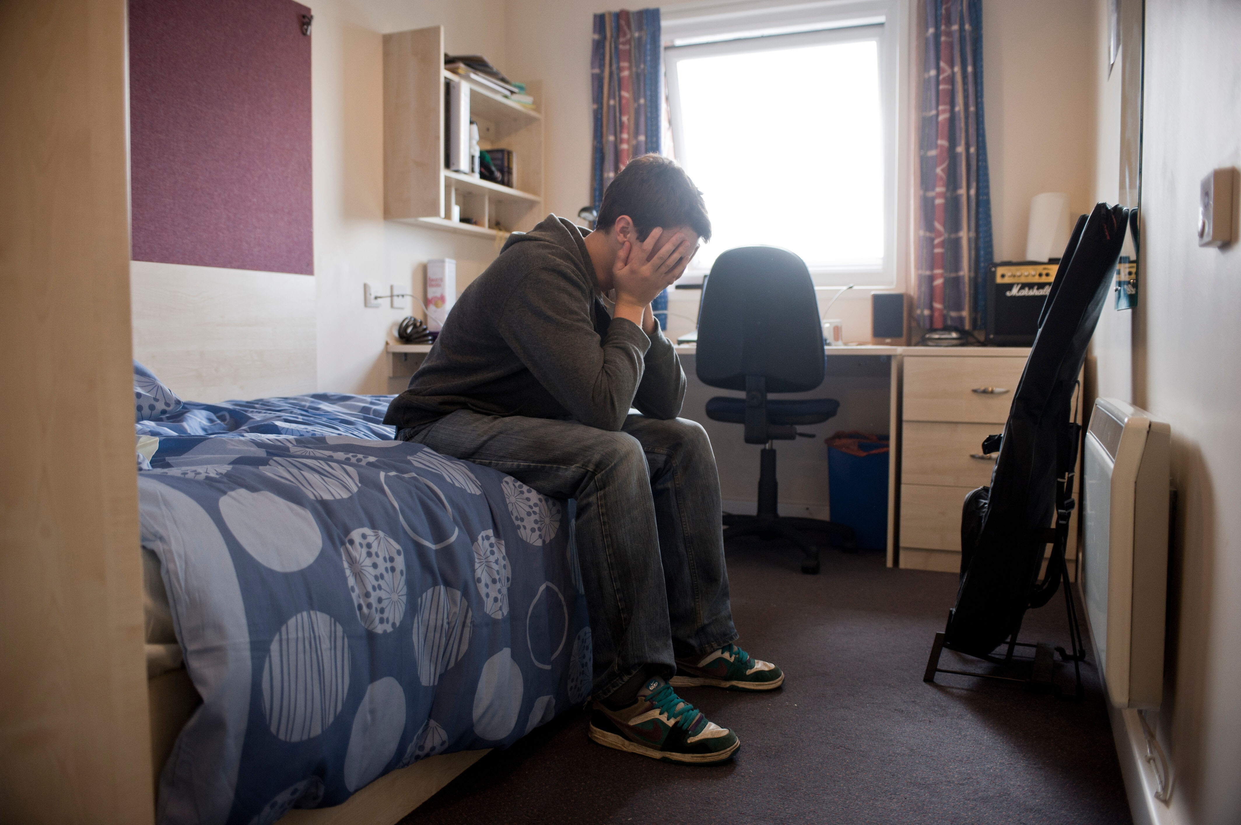 Young Caucasian male student with head in hands, sitting on a bed, alone in room away from home