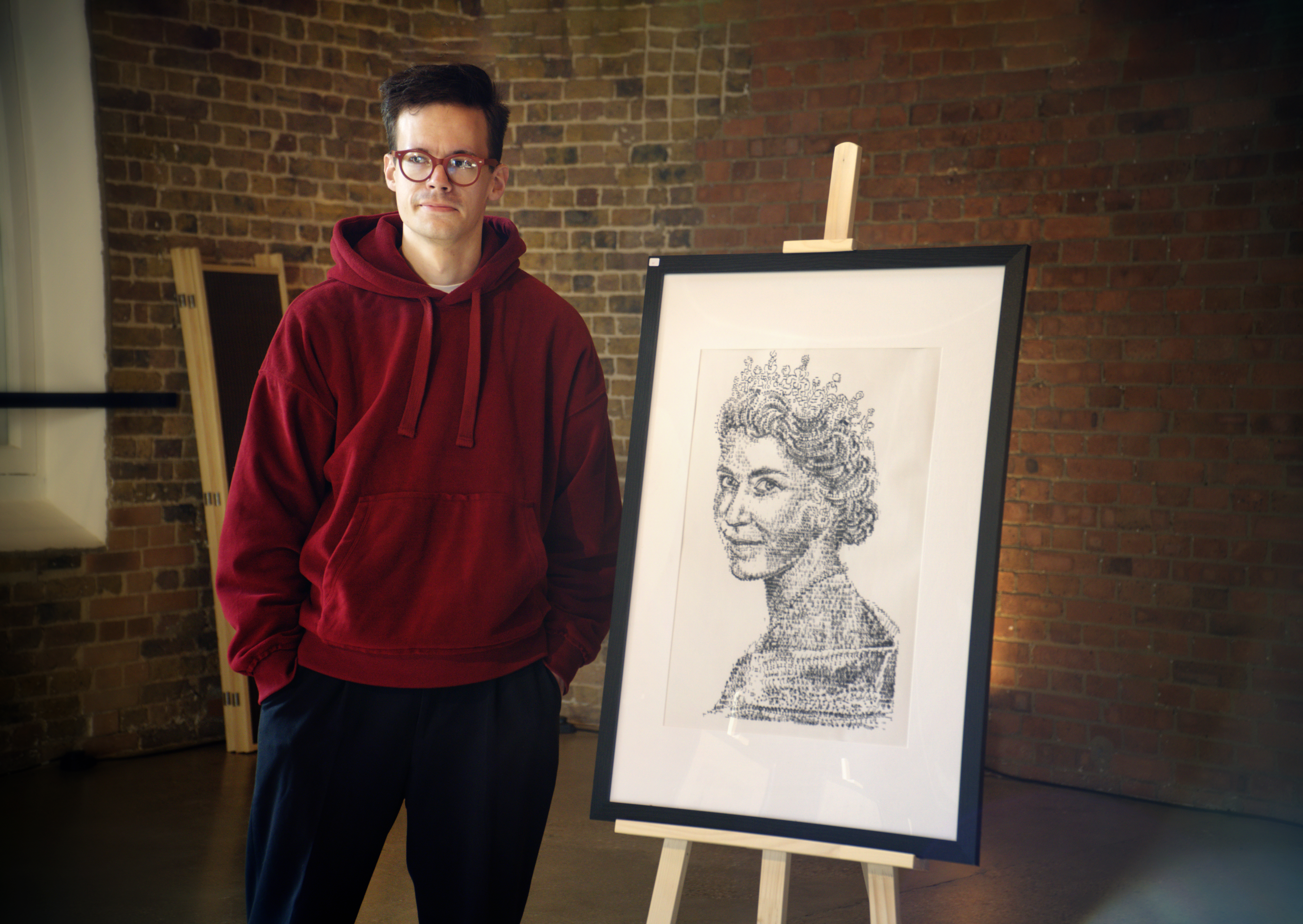 Man in red hoodie and glasses next to a portrait of The Queen 