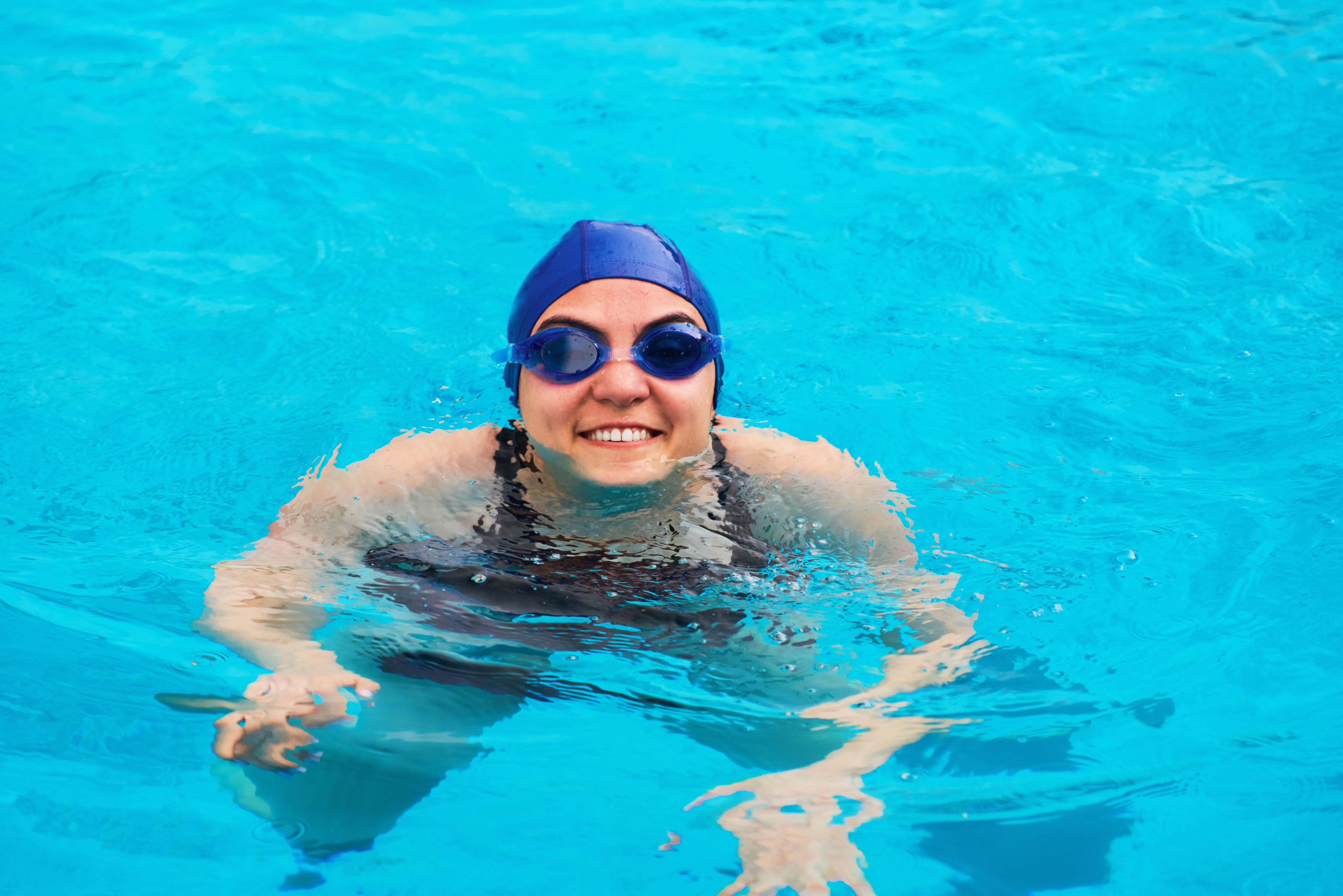 Woman in a swimming pool, smiling at the camera, wearing goggles and a blue swim cap
