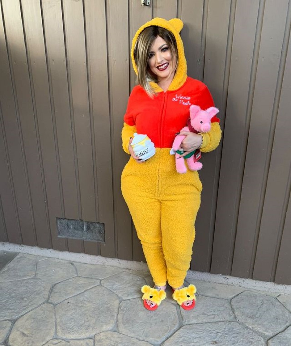 Woman wearing yellow and red onesie and holding a stuffed pink pig toy and a honey jar in her hands