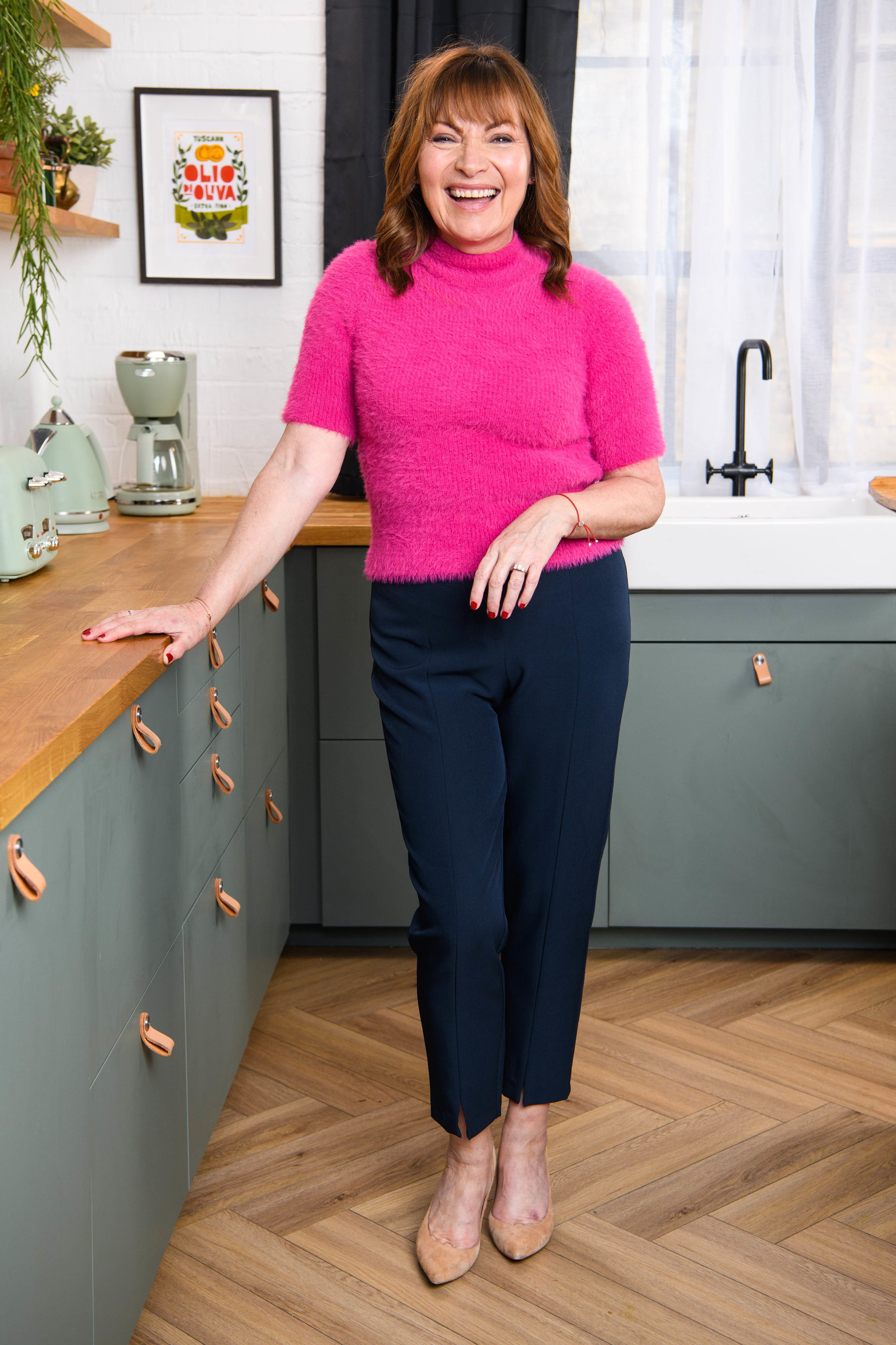 Lorraine Kelly in a pink top and blue trousers, standing in a kitchen, smiling at the camera