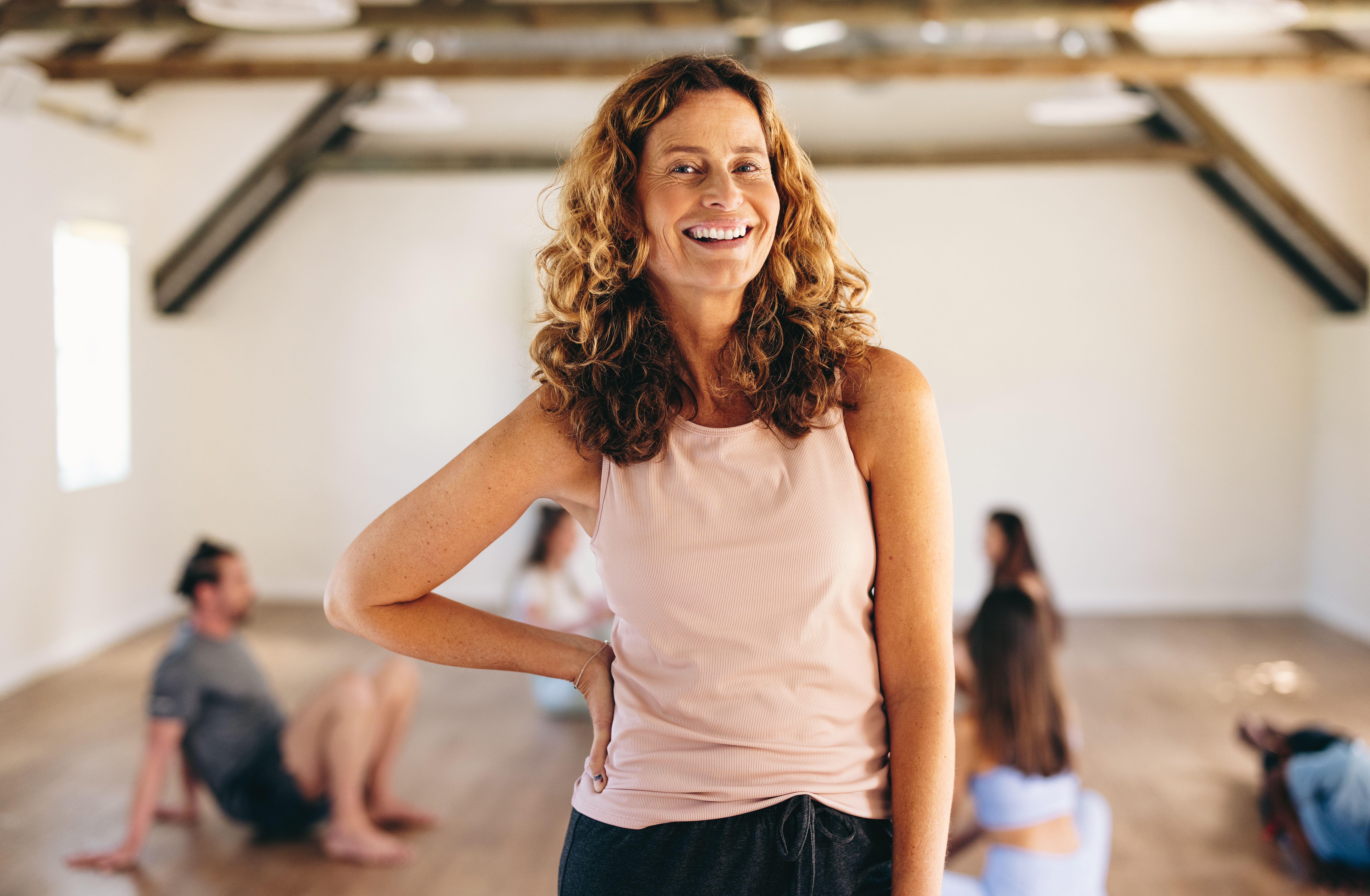 Middle aged white woman at a yoga class, standing up and smiling at the camera