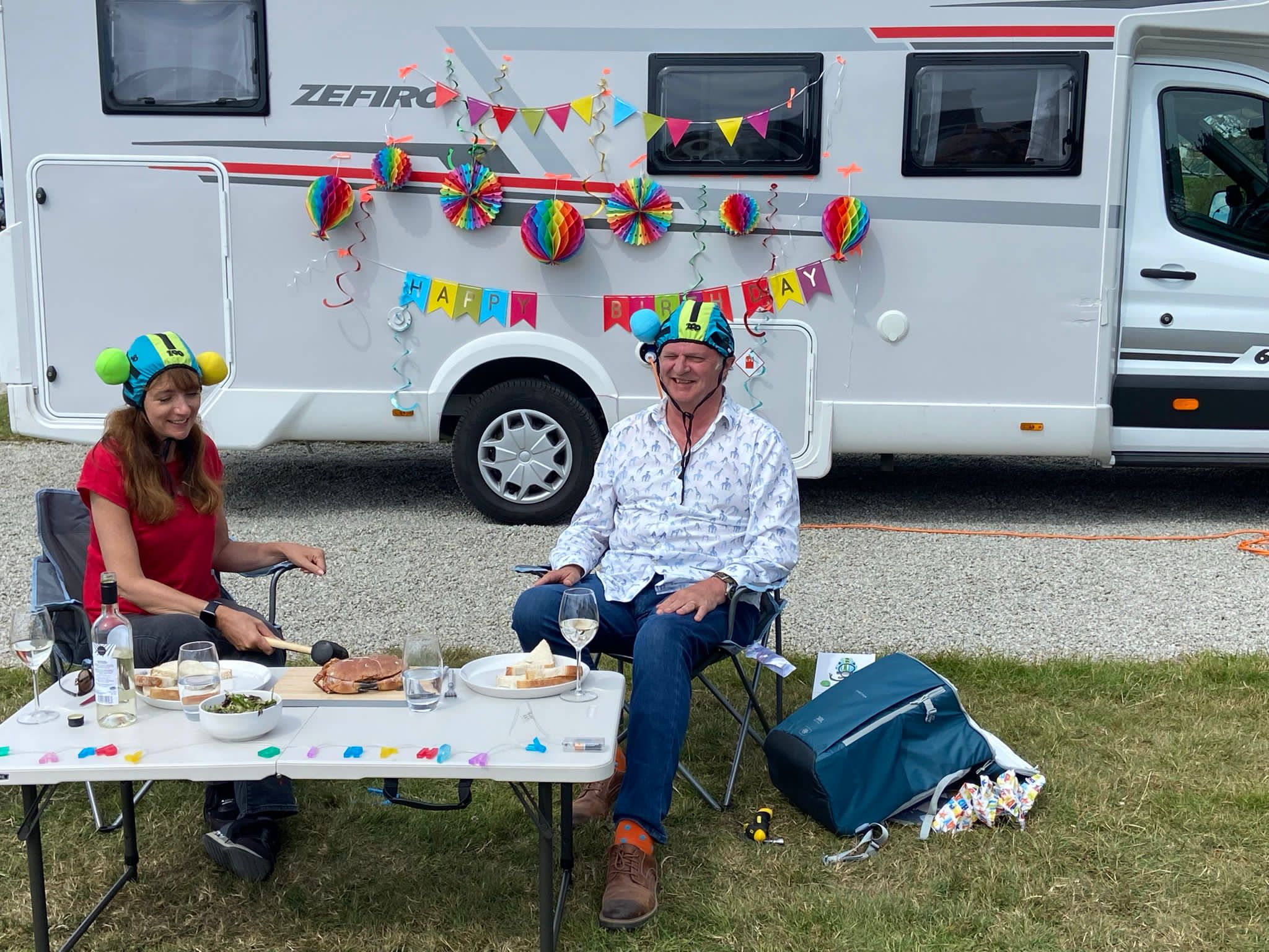 Paul Merton & Suki Webster sitting at a table outside their motorhome with a Happy Birthday banner behind them (Paul Merton & Suki Webster/PA)