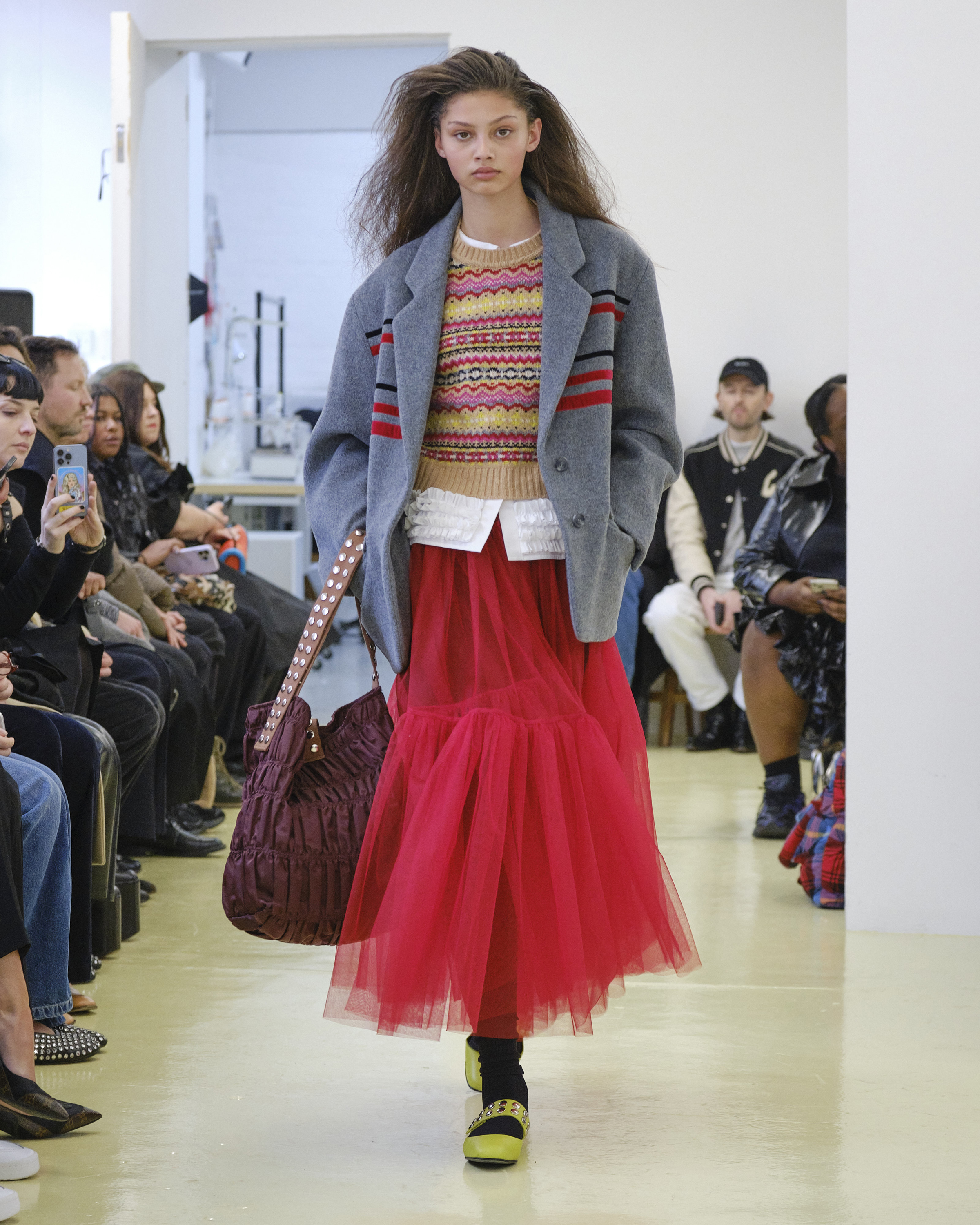 A model on the Molly Goddard runway at AW23 London Fashion Week, wearing a long red tulle skirt with a patterned woollen jumper and grey jacket, carrying an oversized slouchy bag