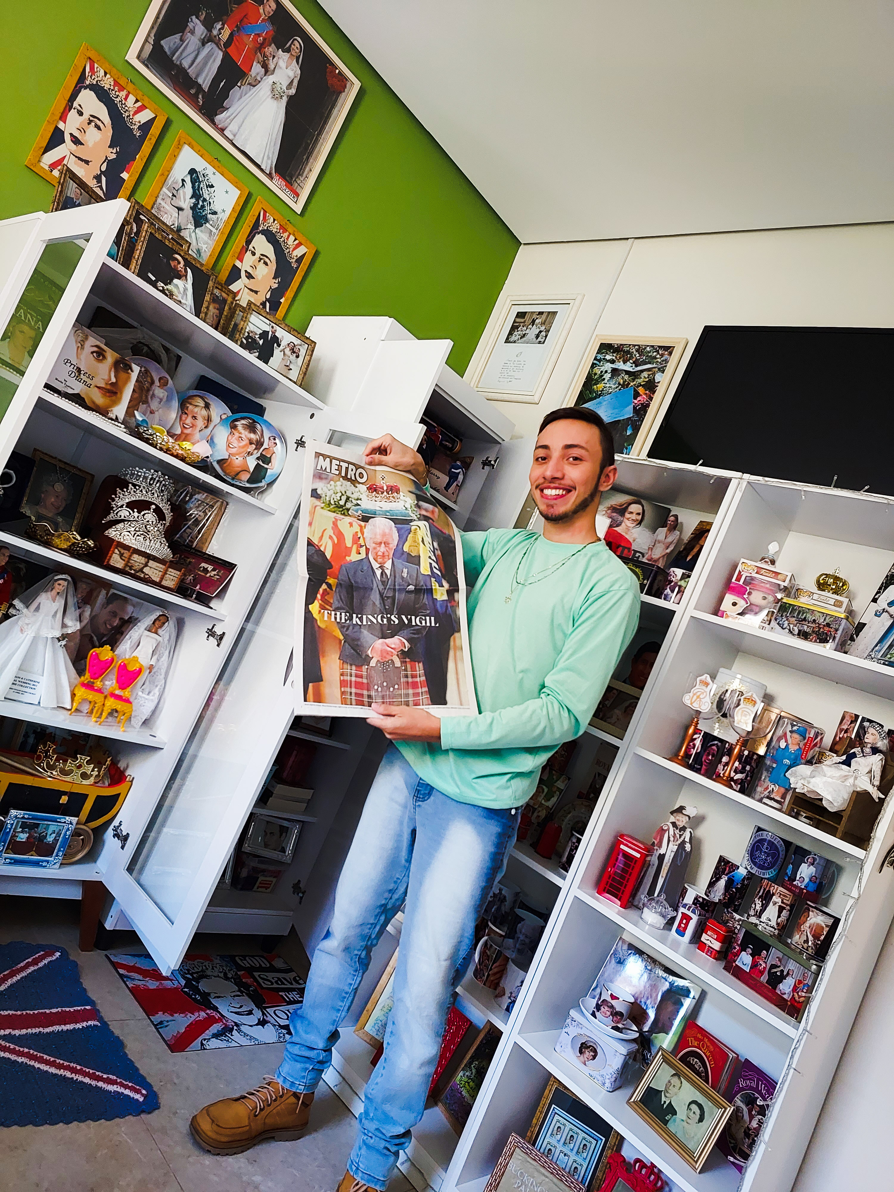 Savio Valei standing in front of his display shelves full of royal memorabilia holding a newspaper with a picture of King Charles III on the front page