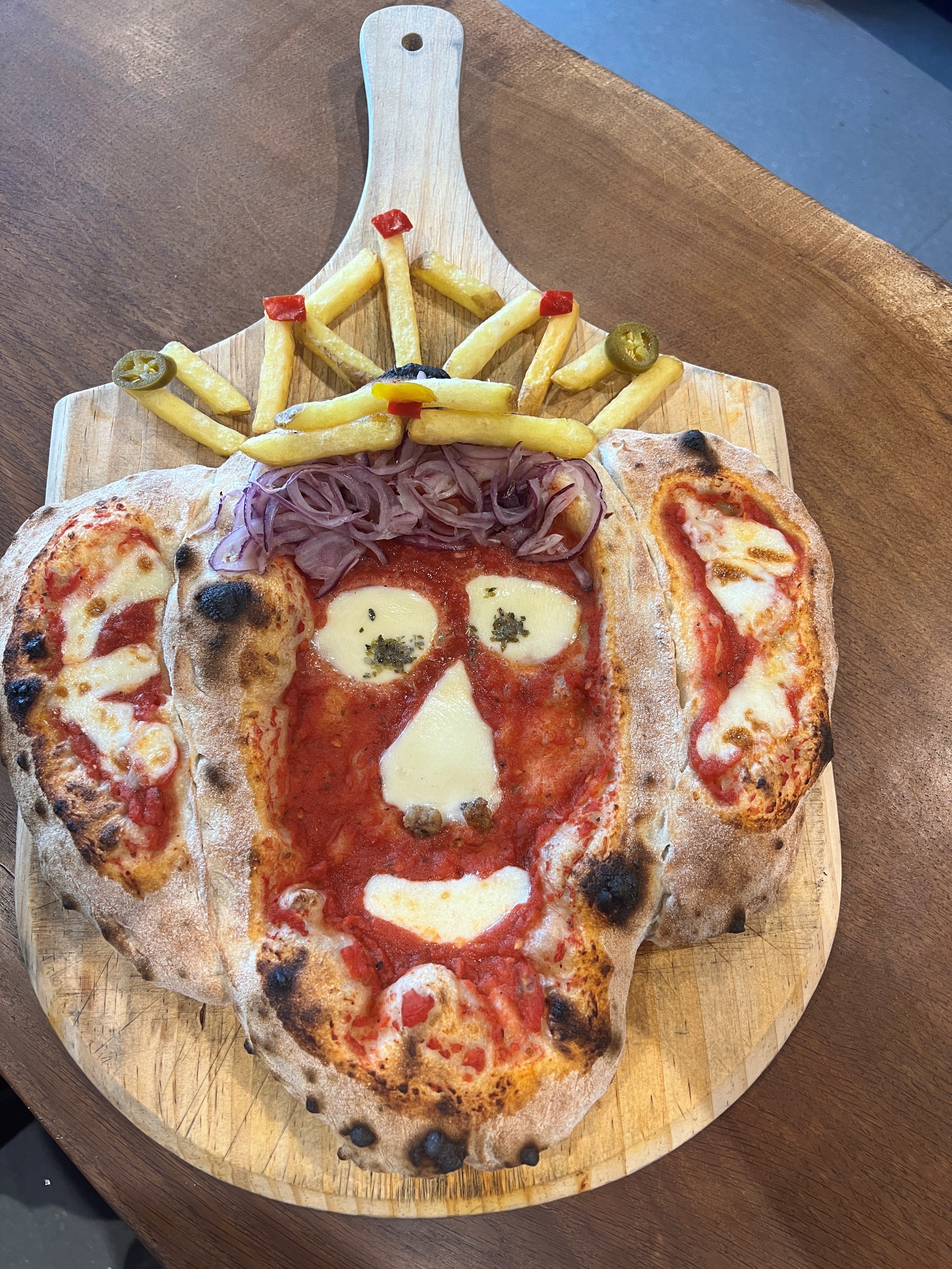 A tomato and mozzarella pizza in the shape of King Charles' head topped with a crown made out of chips, red peppers and jalapenos served on a wooden pizza board