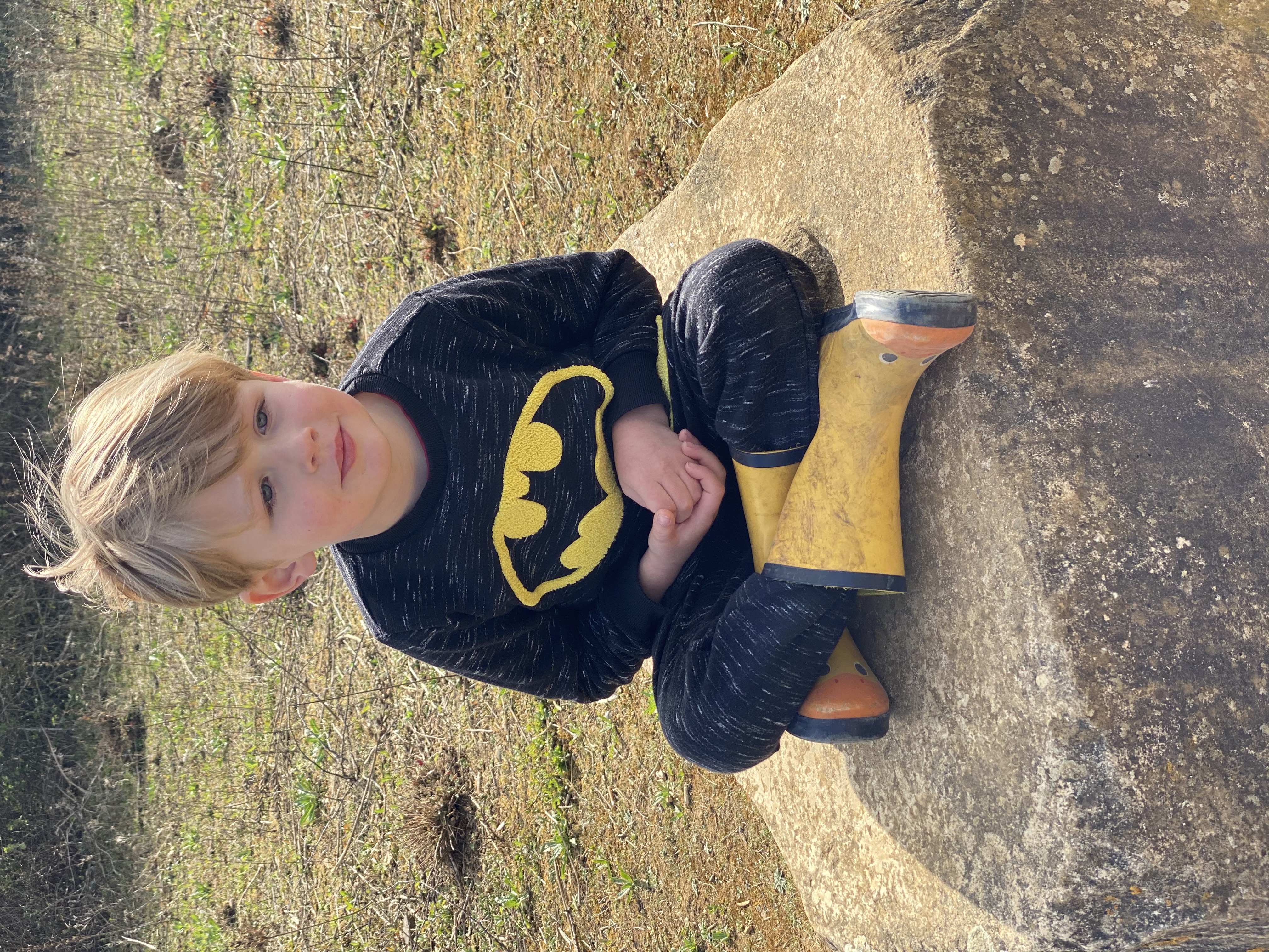 Young boy sat cross-legged on a rock wearing yellow wellies and a Batman top