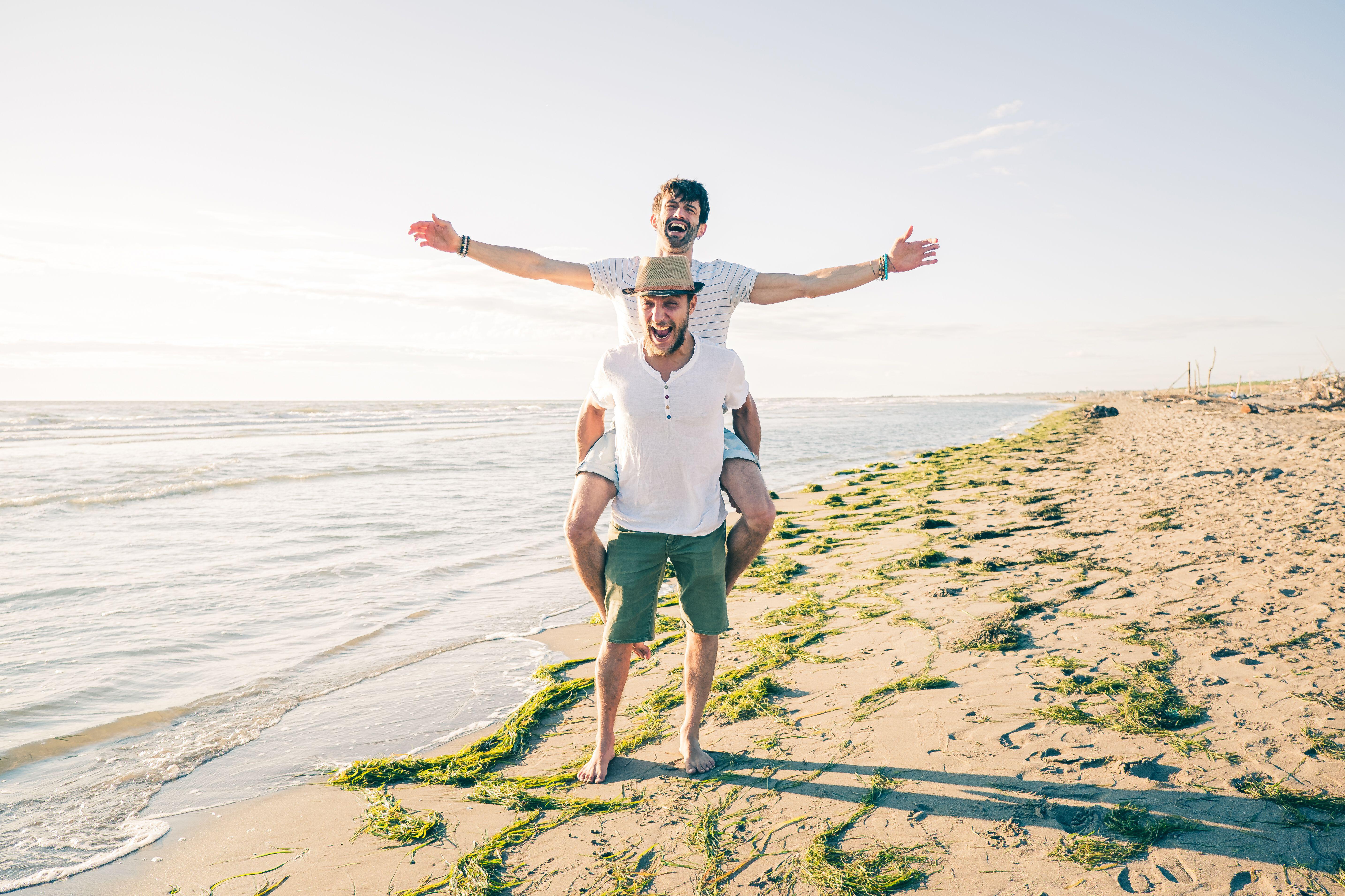 Gay male couple on a beach, one giving the other a piggy back, looking happy and relaxed