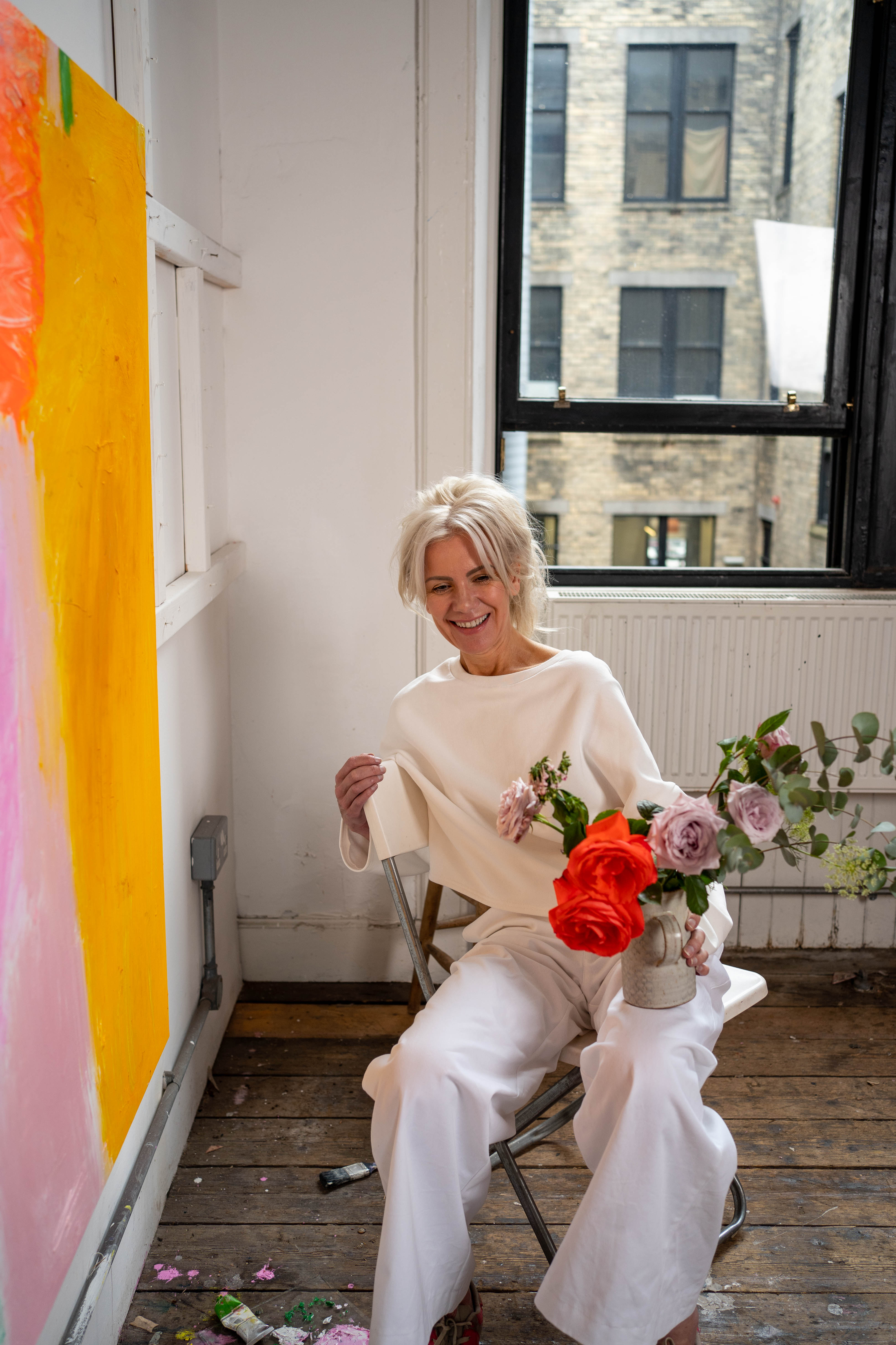 Alison McWhirter dressed in white holding a vase of flowers in front of one of her paintings