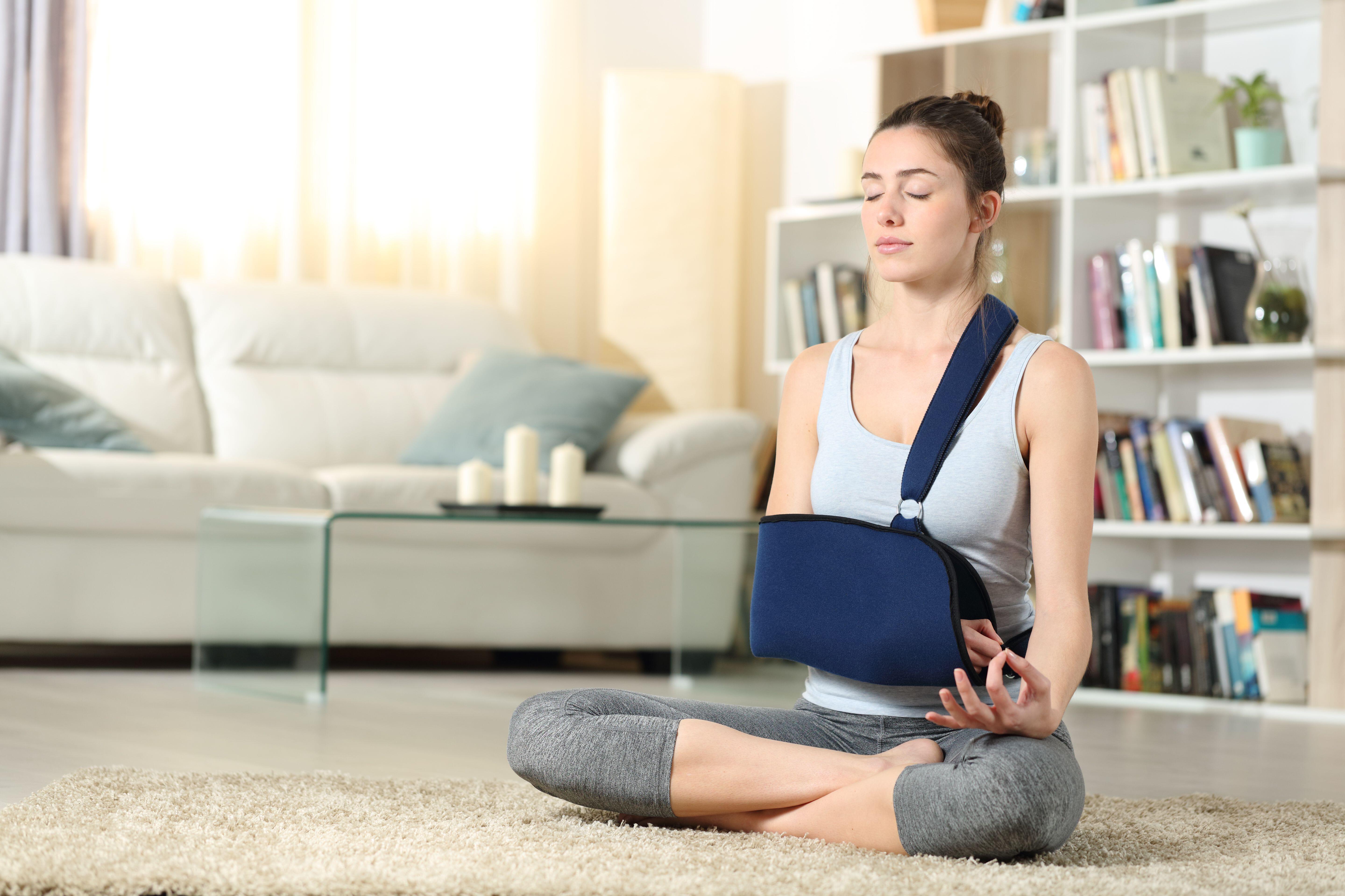 A young woman meditating on the floor at home, with one arm in a joint support sling