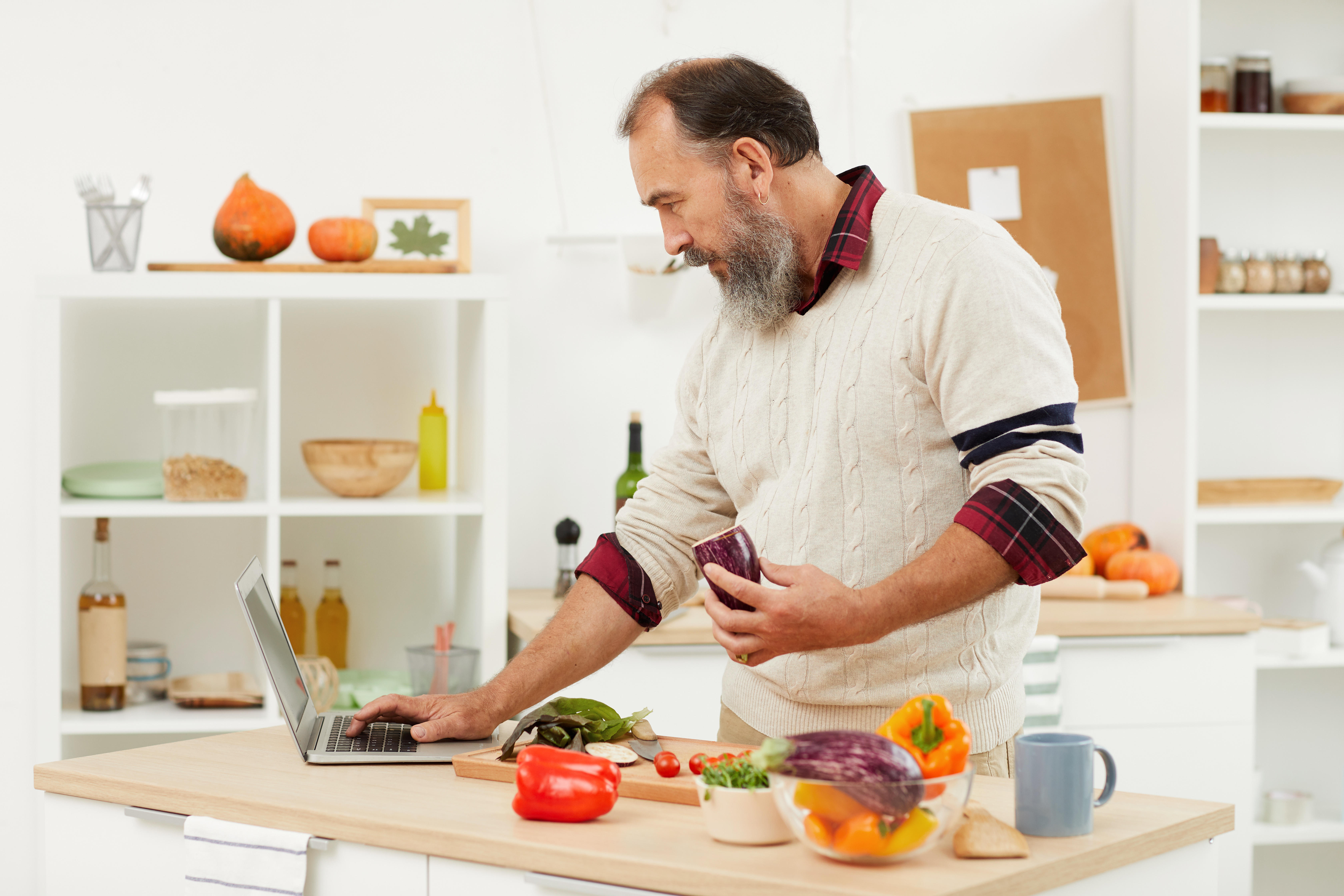 A middle aged man cooking with veg in his kitchen, looking at a recipe on a laptop