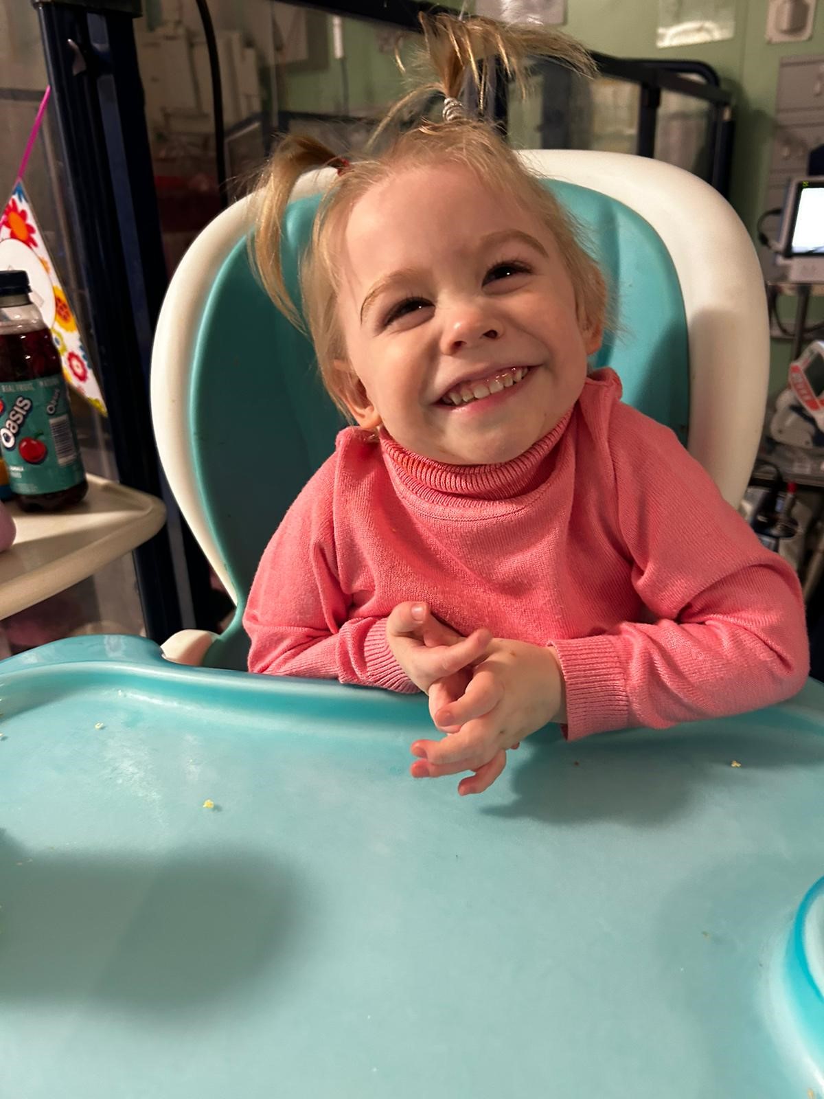 Amelia sitting in a high chair wearing a pink top and smiling at the camera 