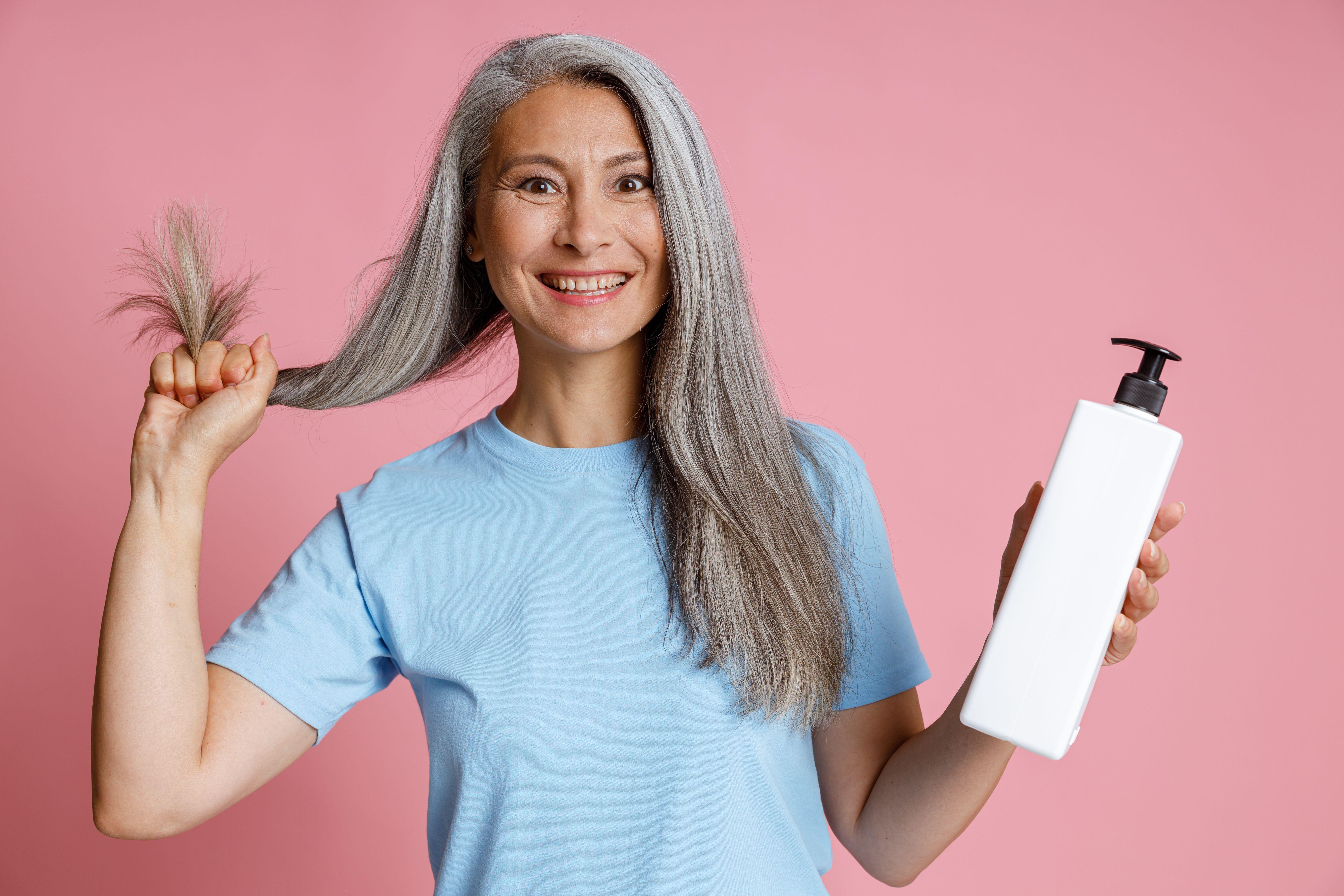 Woman with long grey hair holding a bottle of hair product, standing against a pink background, looking happy and confident