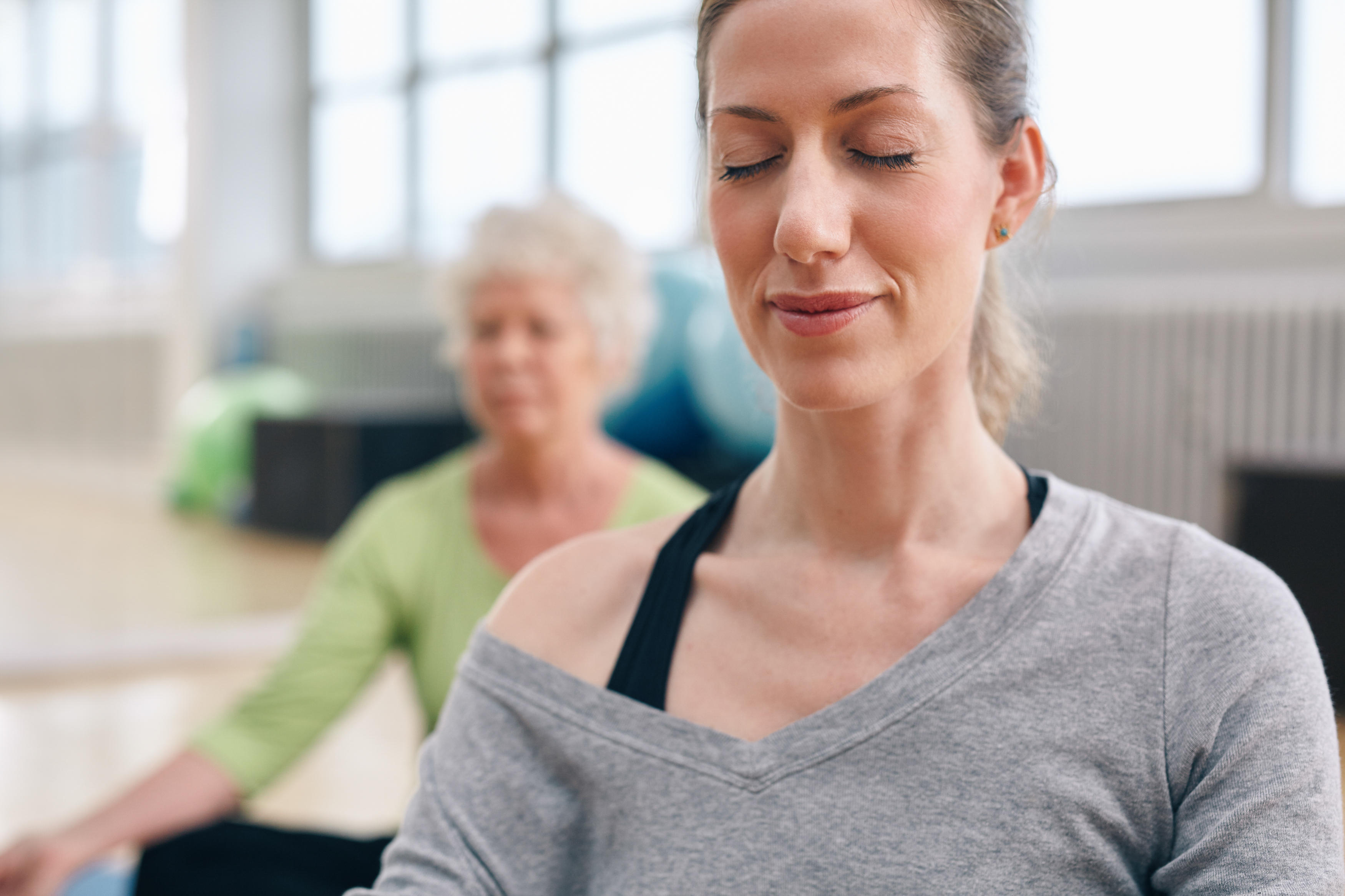 Relaxed woman practicing yoga in gym with senior woman in background meditating
