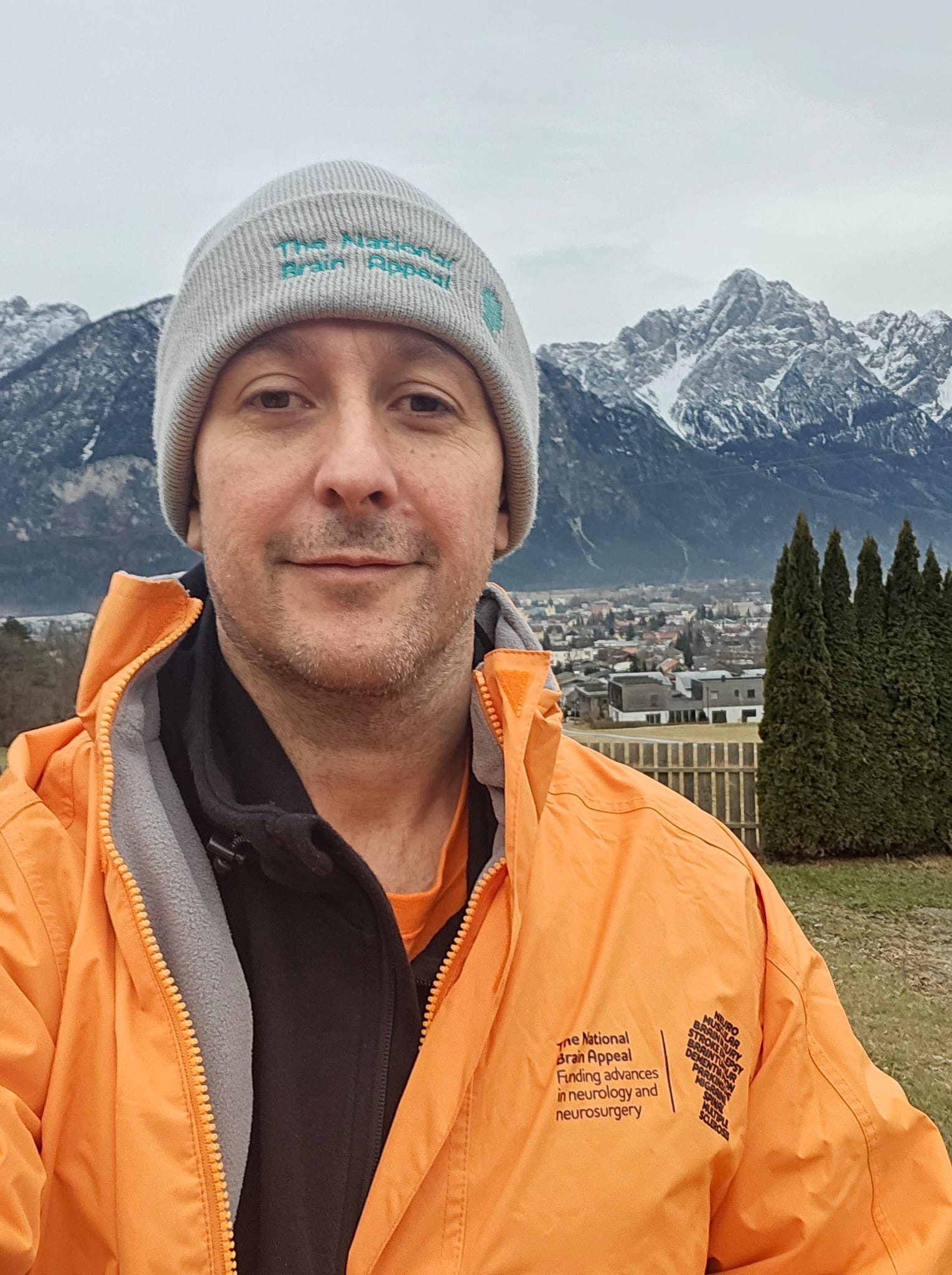 A photo of James Dick wearing an orange jacket and a hat with a mountain range behind him