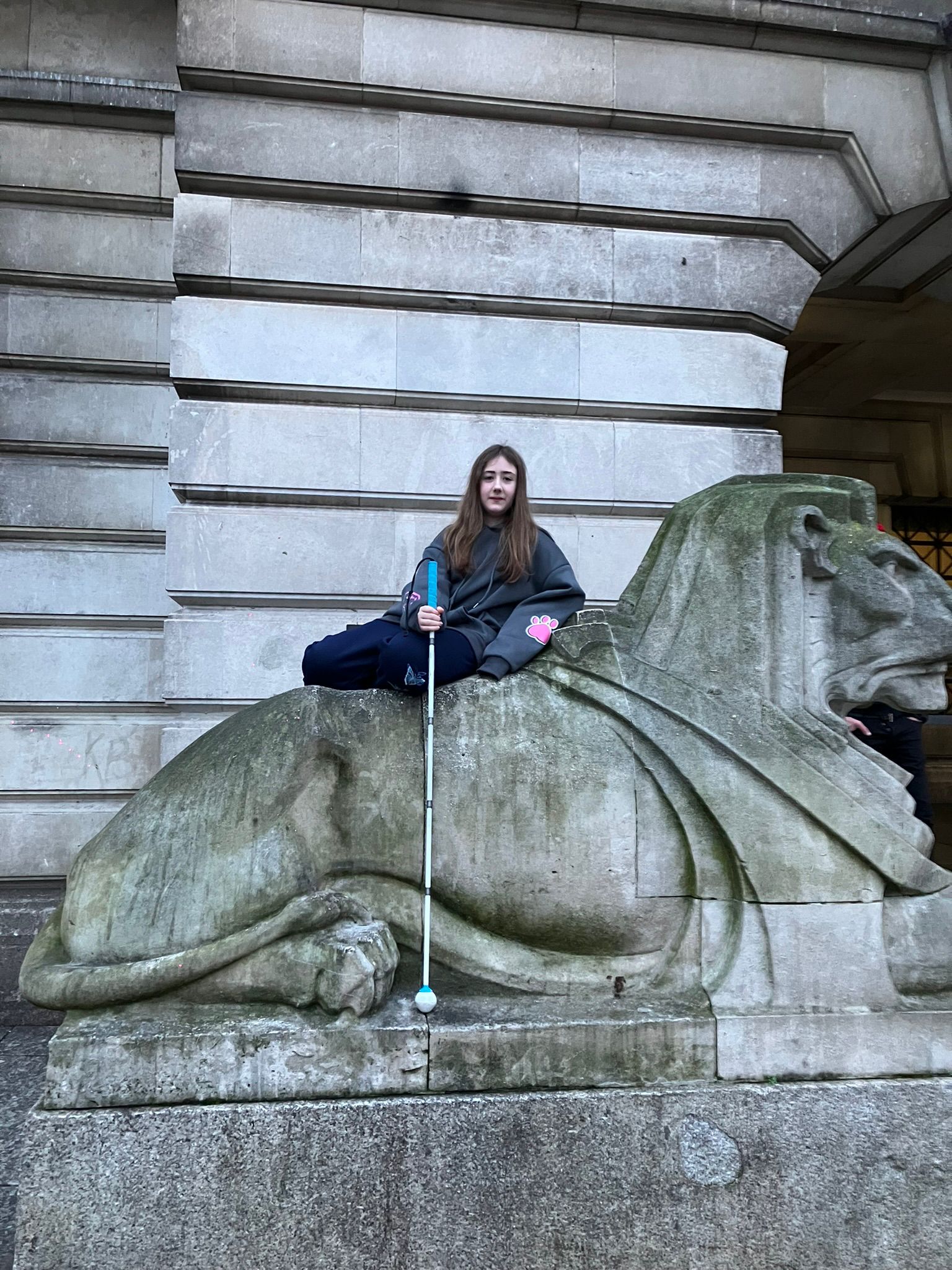 Visually impaired girl holding a cane whilst sat on top of a lion statue