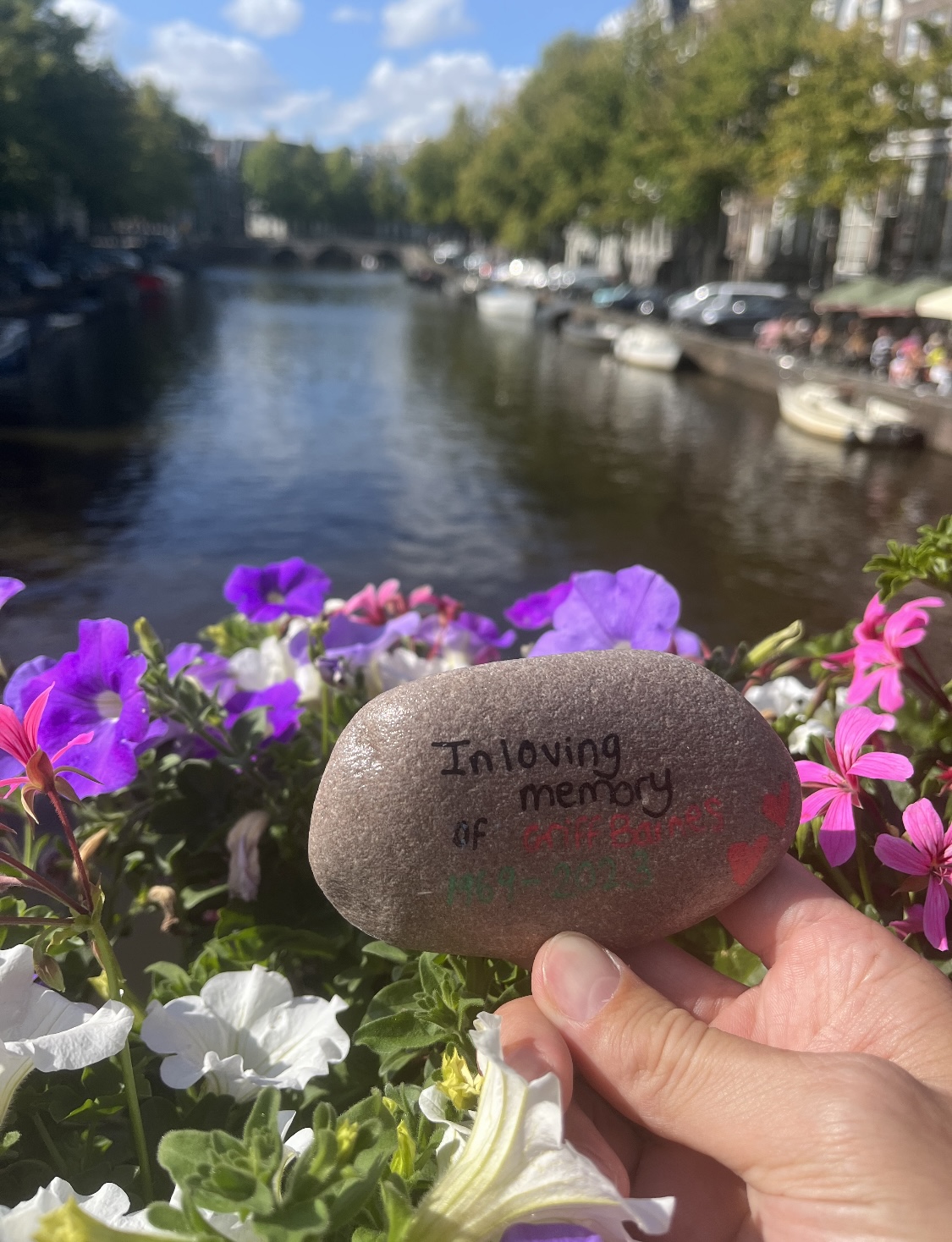 A rock reading the words 'In loving memory of Griff Barnes' held in front of some flowers over a canal in Amsterdam 