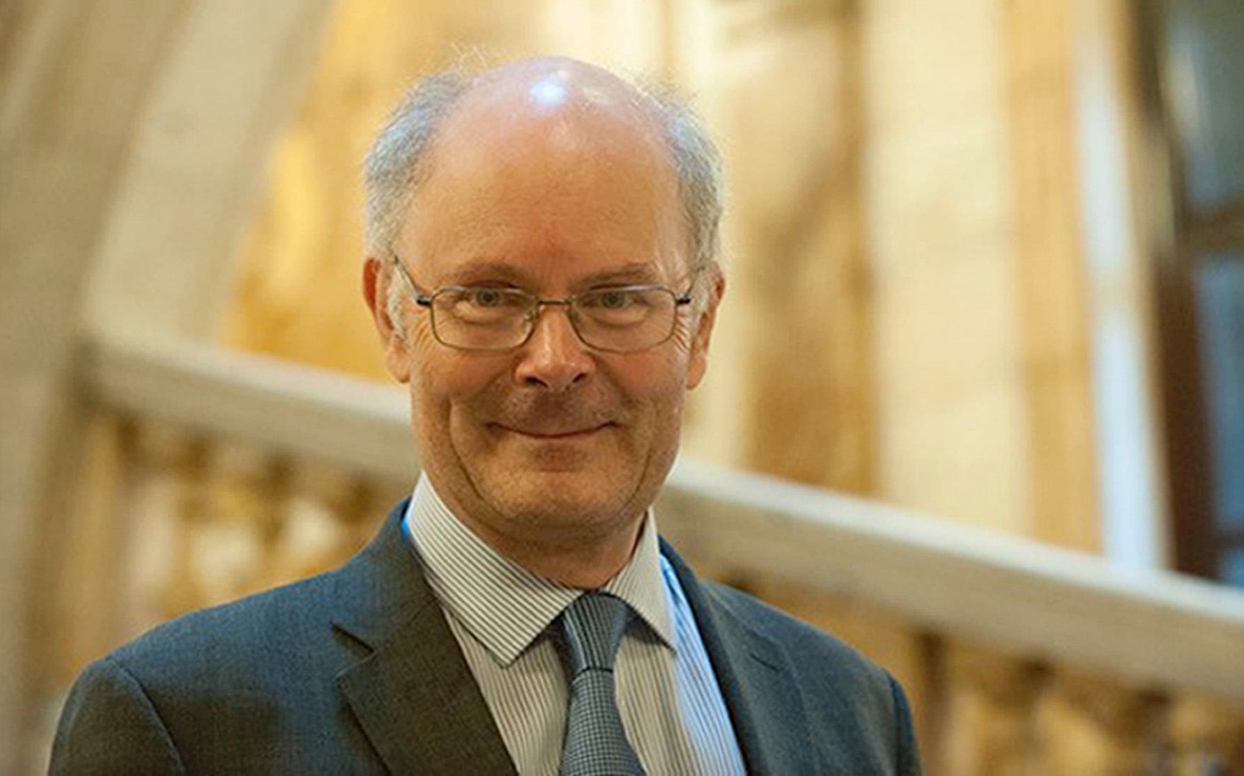 Professor Sir John Curtice smiling, pictured in front of a staircase