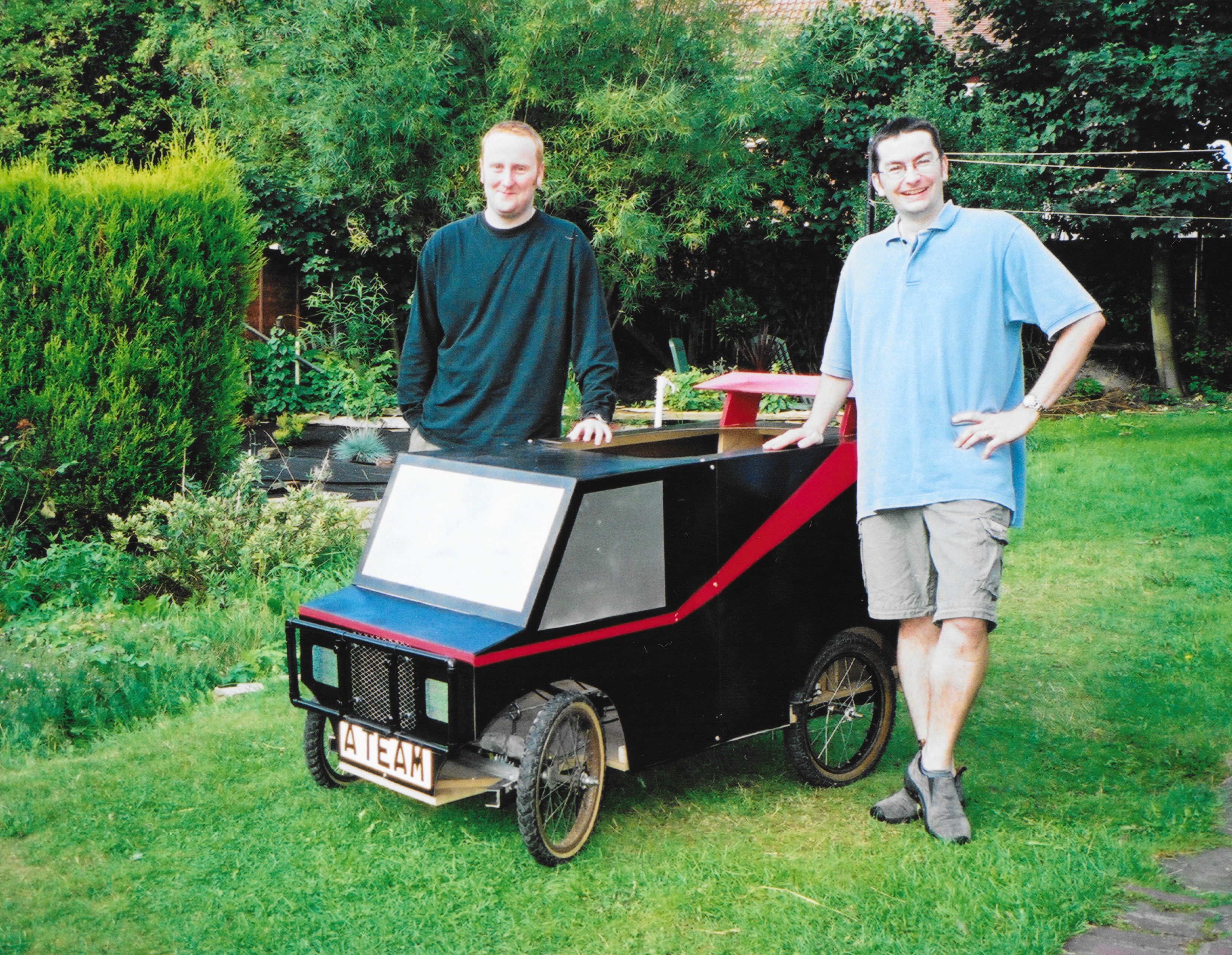 Two men standing next to a soapbox vehicle in a garden